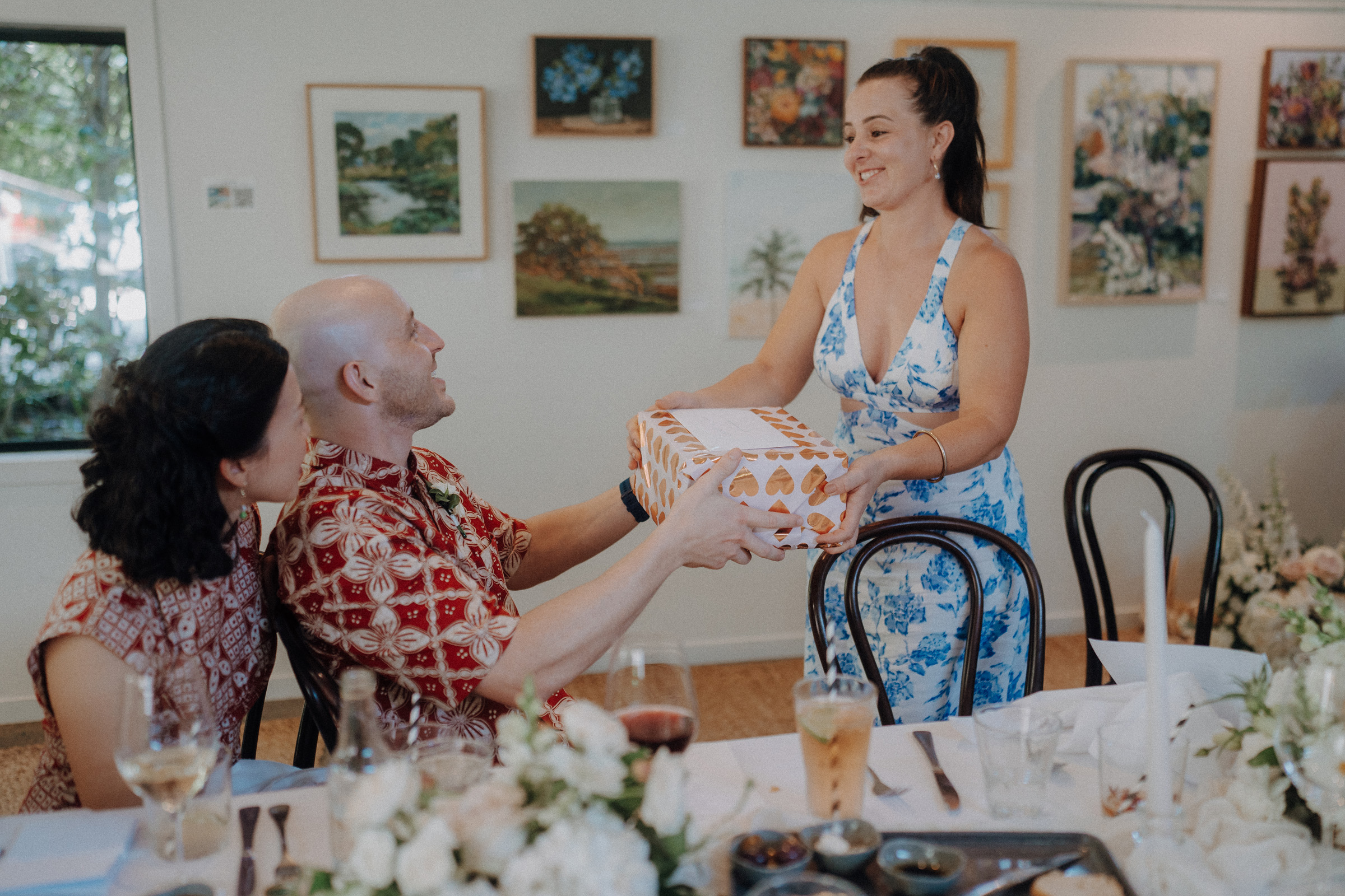 A woman in a floral dress hands a wrapped gift to a seated man wearing a red patterned shirt, while another woman looks on. They are in a room with wall art and a decorated dining table.