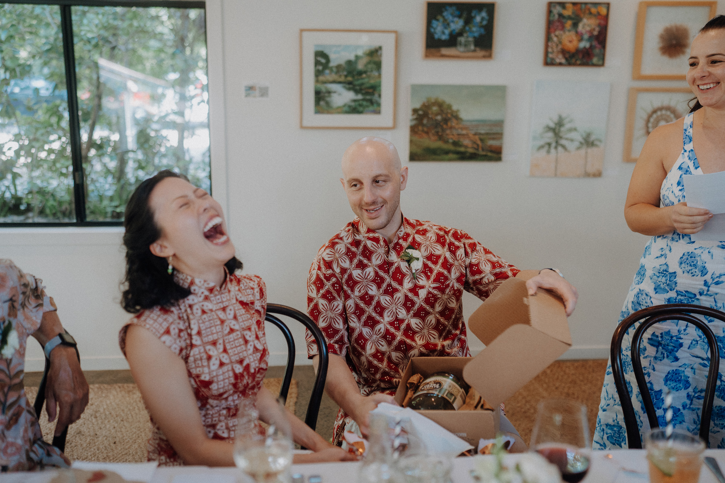 Person laughing while another opens a gift box at a dining table, with wall art and a window in the background.