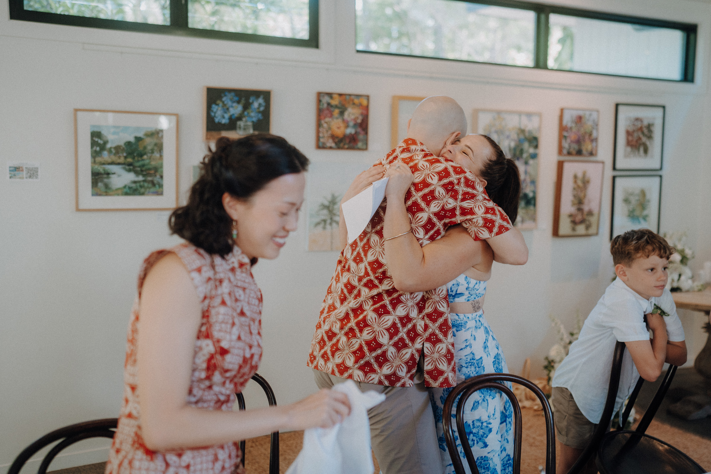 Three people are socializing at an art gallery; two are embracing, and one is standing nearby. Artworks are displayed on the wall behind them. A seated child is also visible.