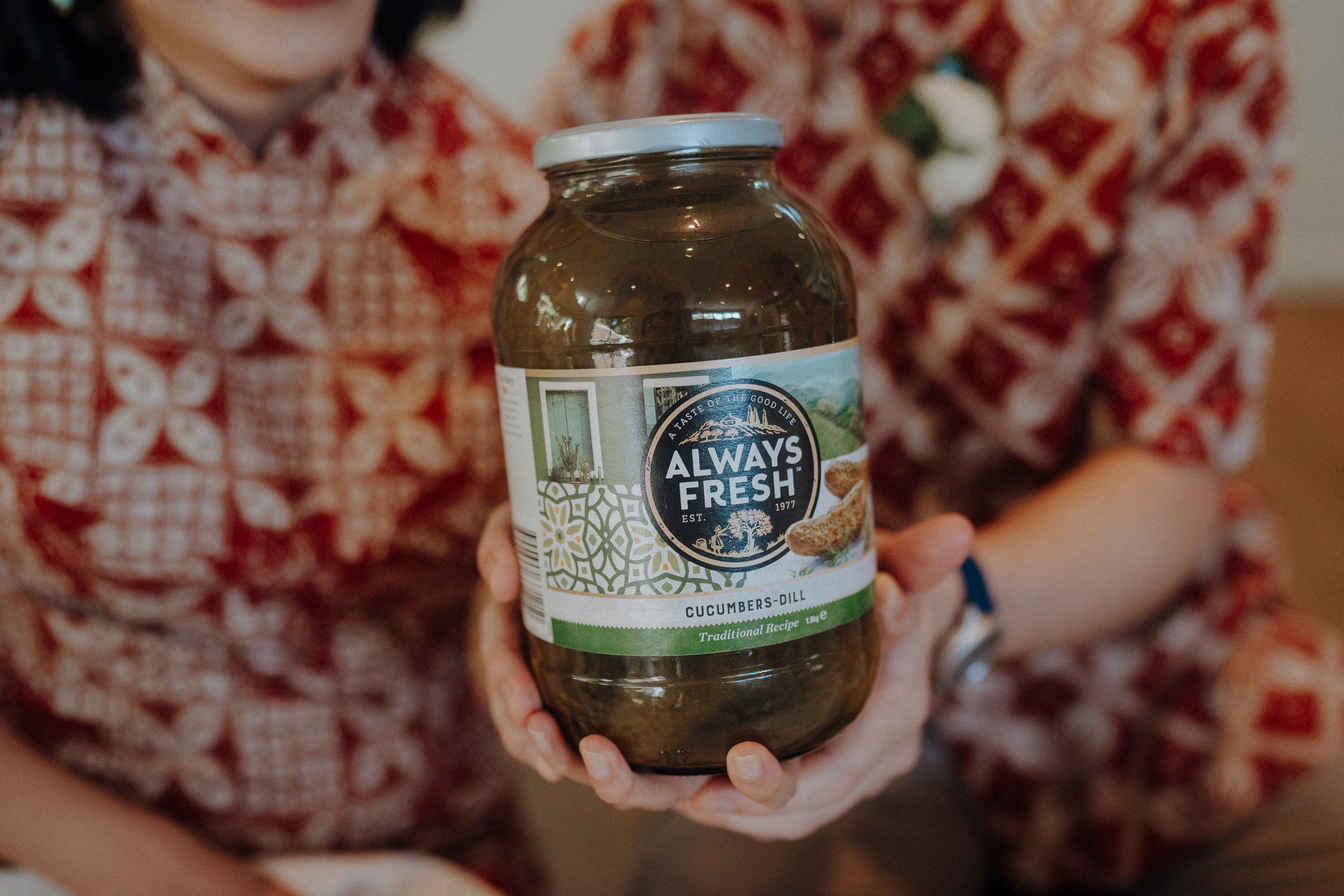 Two people hold a jar labeled "Always Fresh Cucumbers Dill" with patterned shirts in the background.