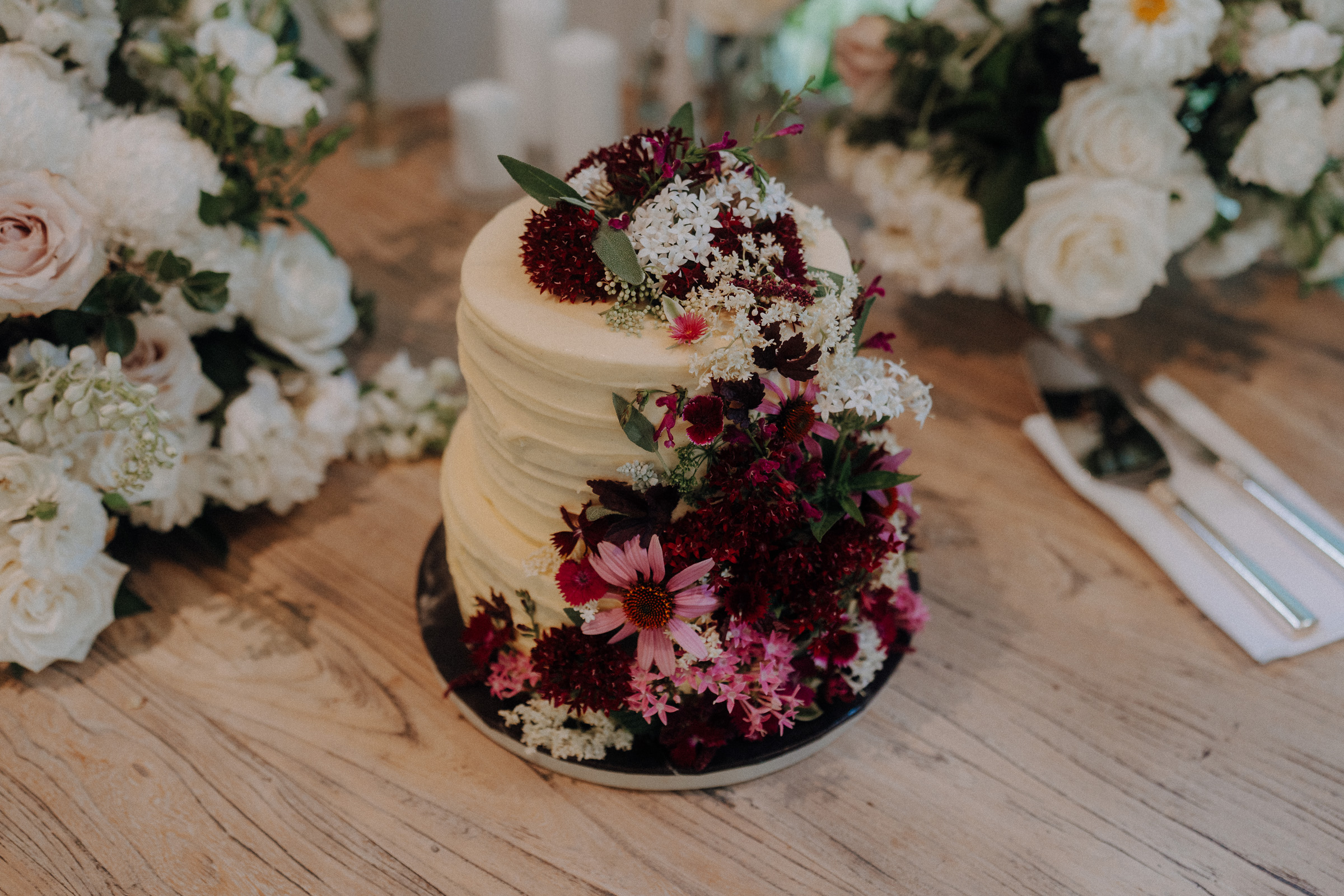 A small, rustic layered cake decorated with red, pink, and white flowers, surrounded by floral arrangements on a wooden table.