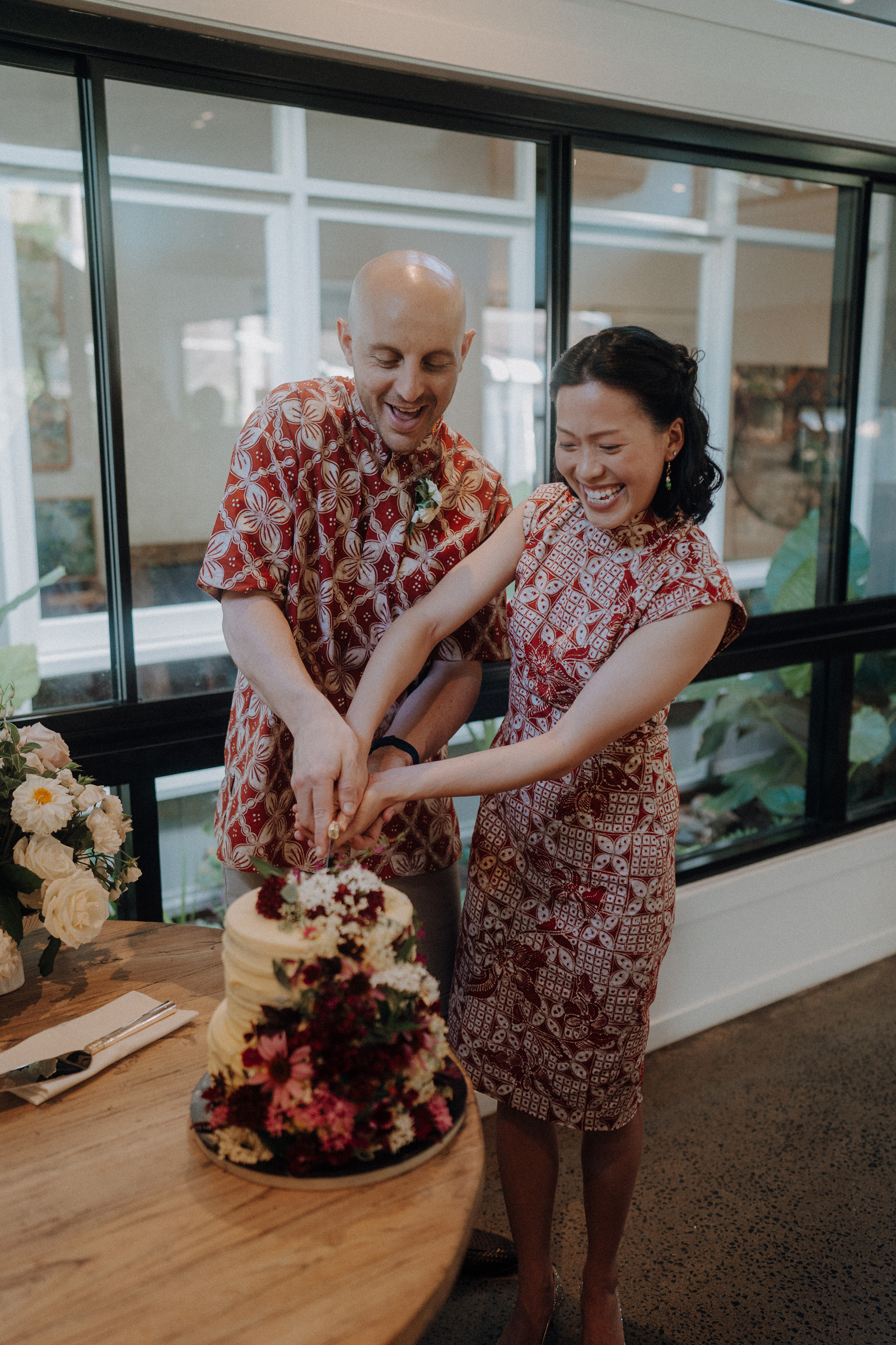 Couple in matching patterned outfits cut a white wedding cake with flowers, standing by a window.