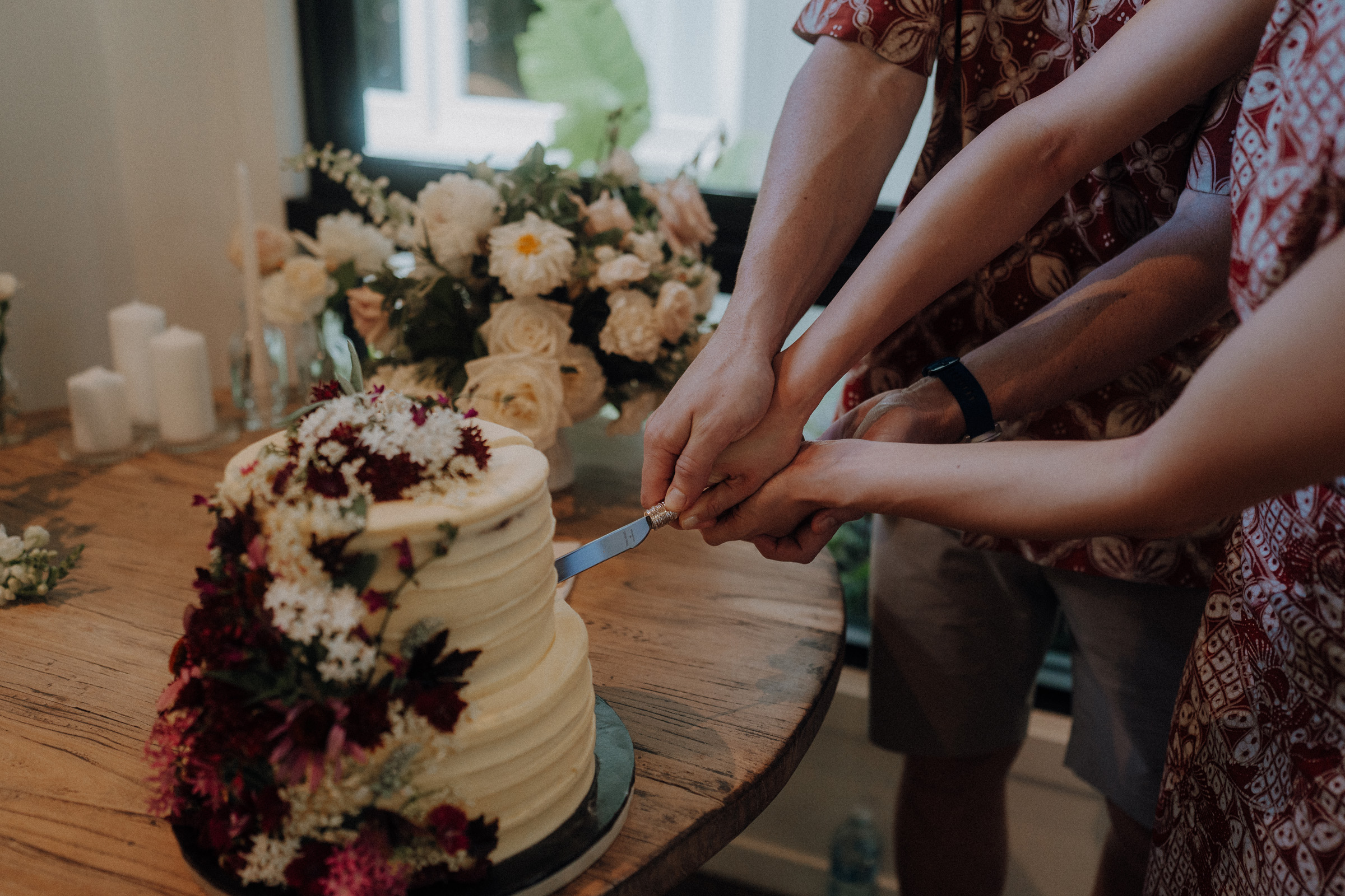 Two people in patterned shirts cut a floral-decorated cake together on a wooden table, with candles and a floral arrangement nearby.