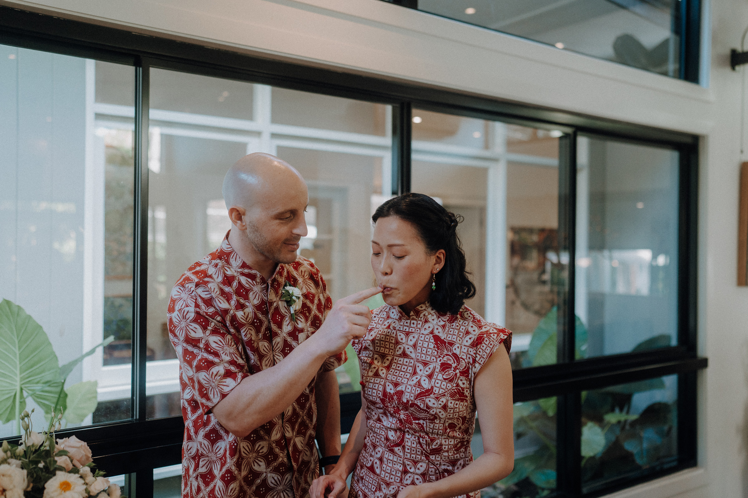 A man in a patterned shirt feeds food to a woman in a matching dress indoors. They stand near a window, surrounded by plants.