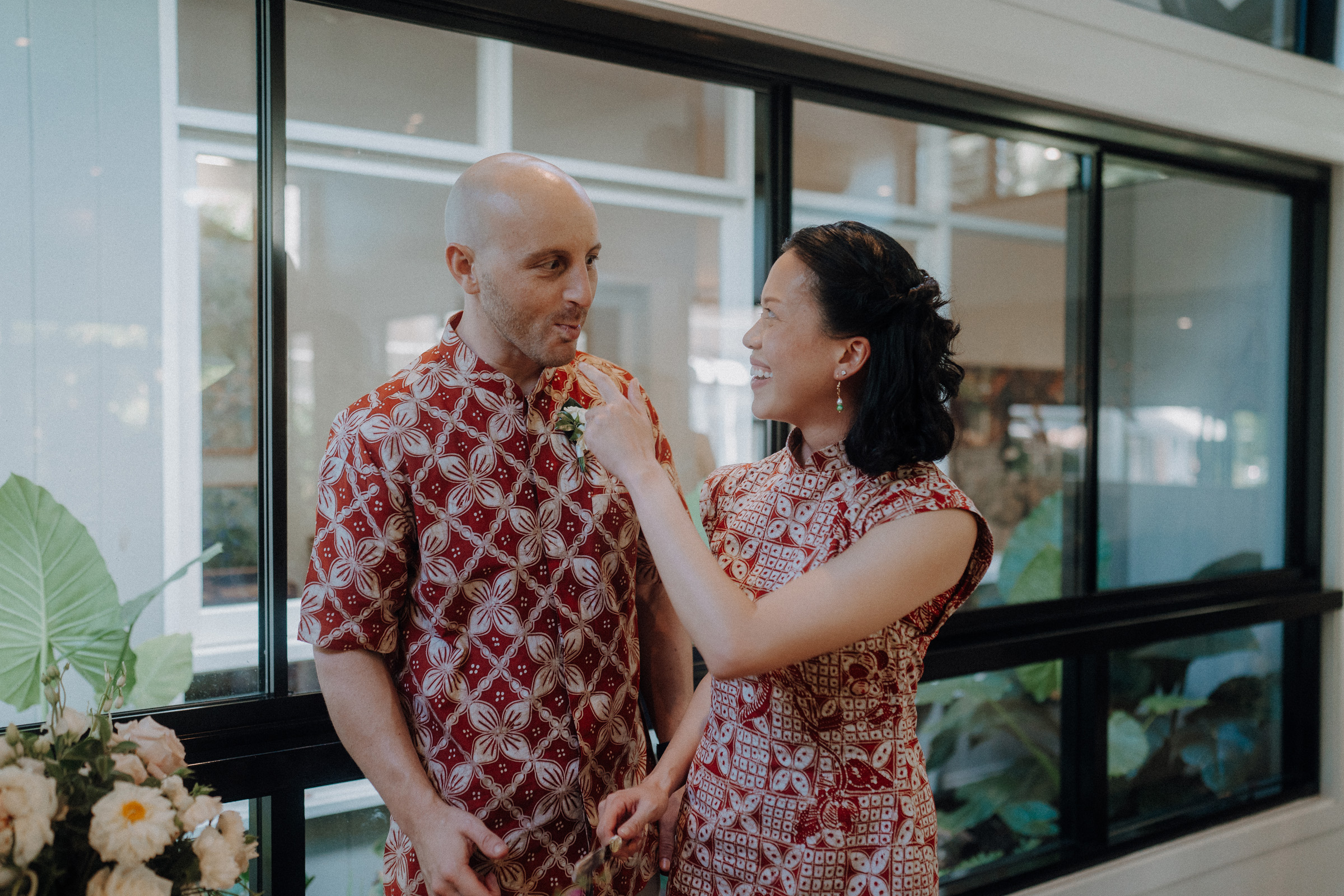 A man and woman in patterned outfits stand indoors near a window. She is smiling and adjusting his shirt.