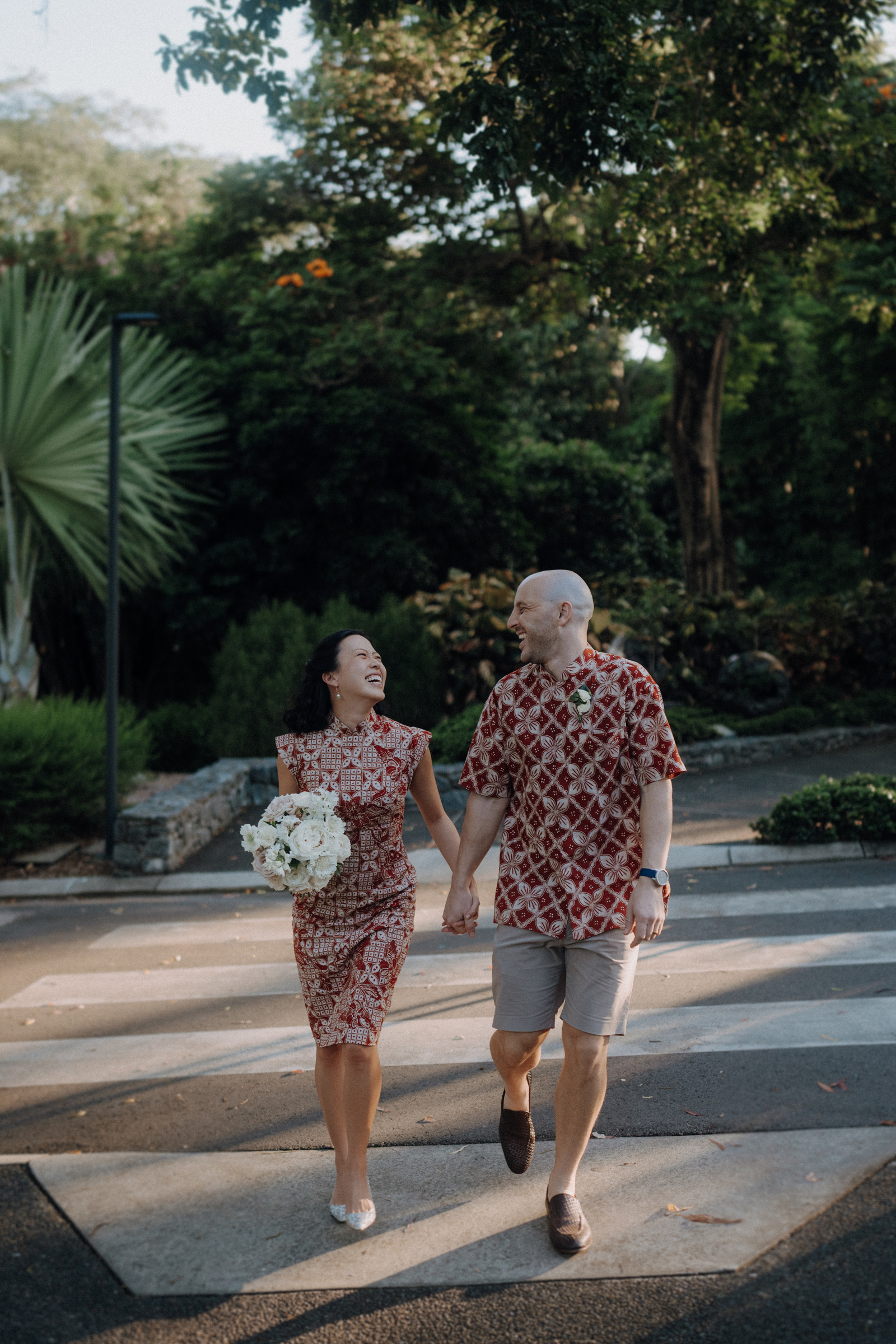 A couple walks hand in hand on a sunny day, wearing matching red patterned outfits. The woman holds a bouquet of white flowers. Lush greenery surrounds them.