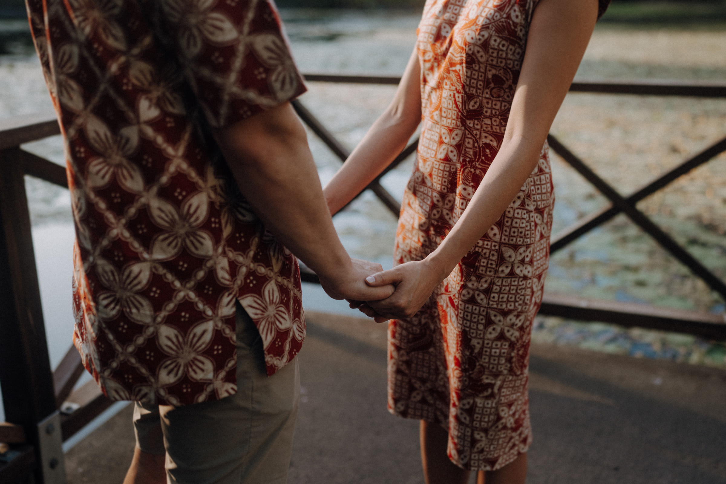 Two people holding hands on a bridge, wearing patterned clothing, with water and lily pads visible in the background.