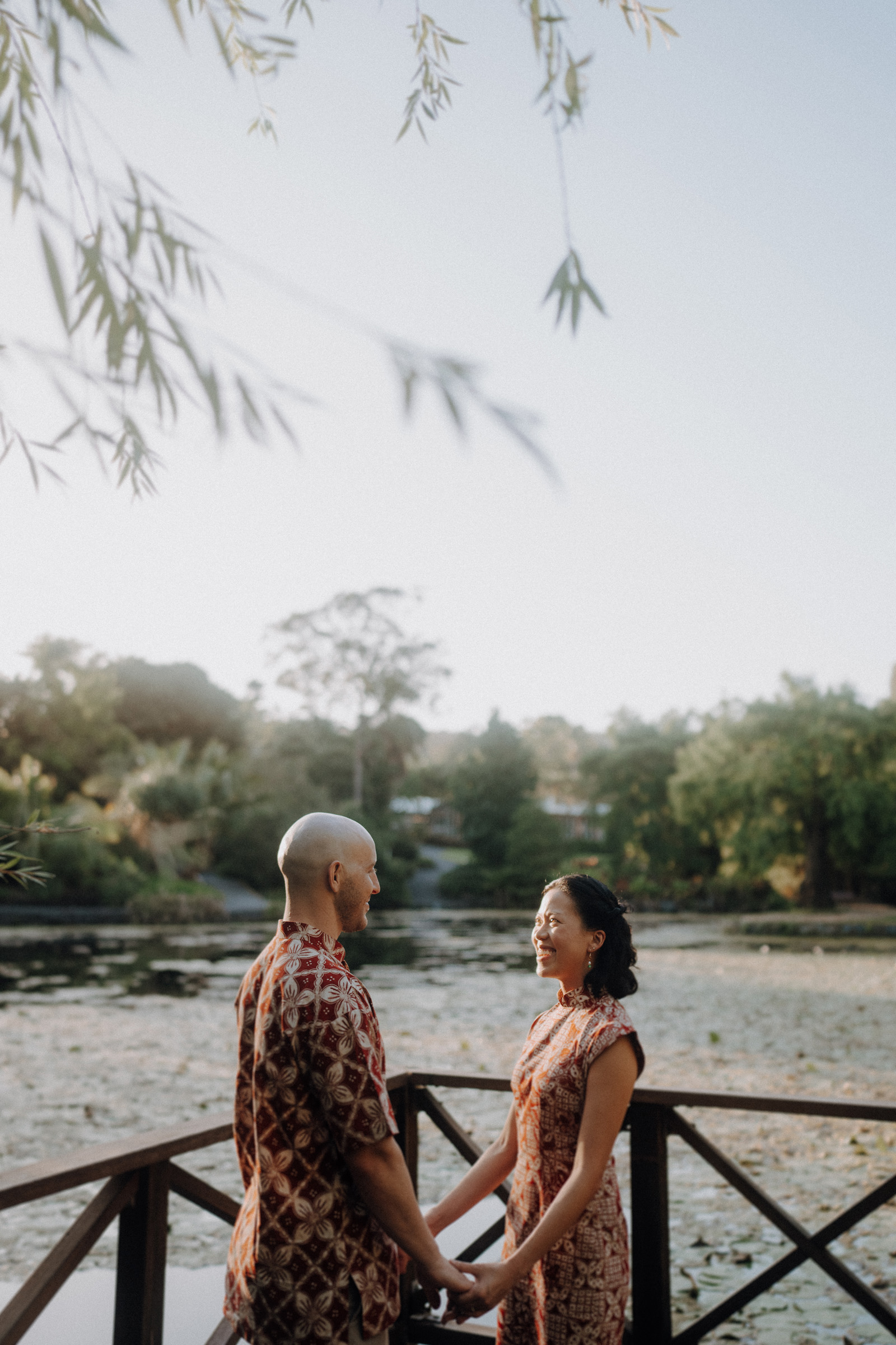 A couple stands on a wooden bridge holding hands, surrounded by greenery and a calm pond in the background.