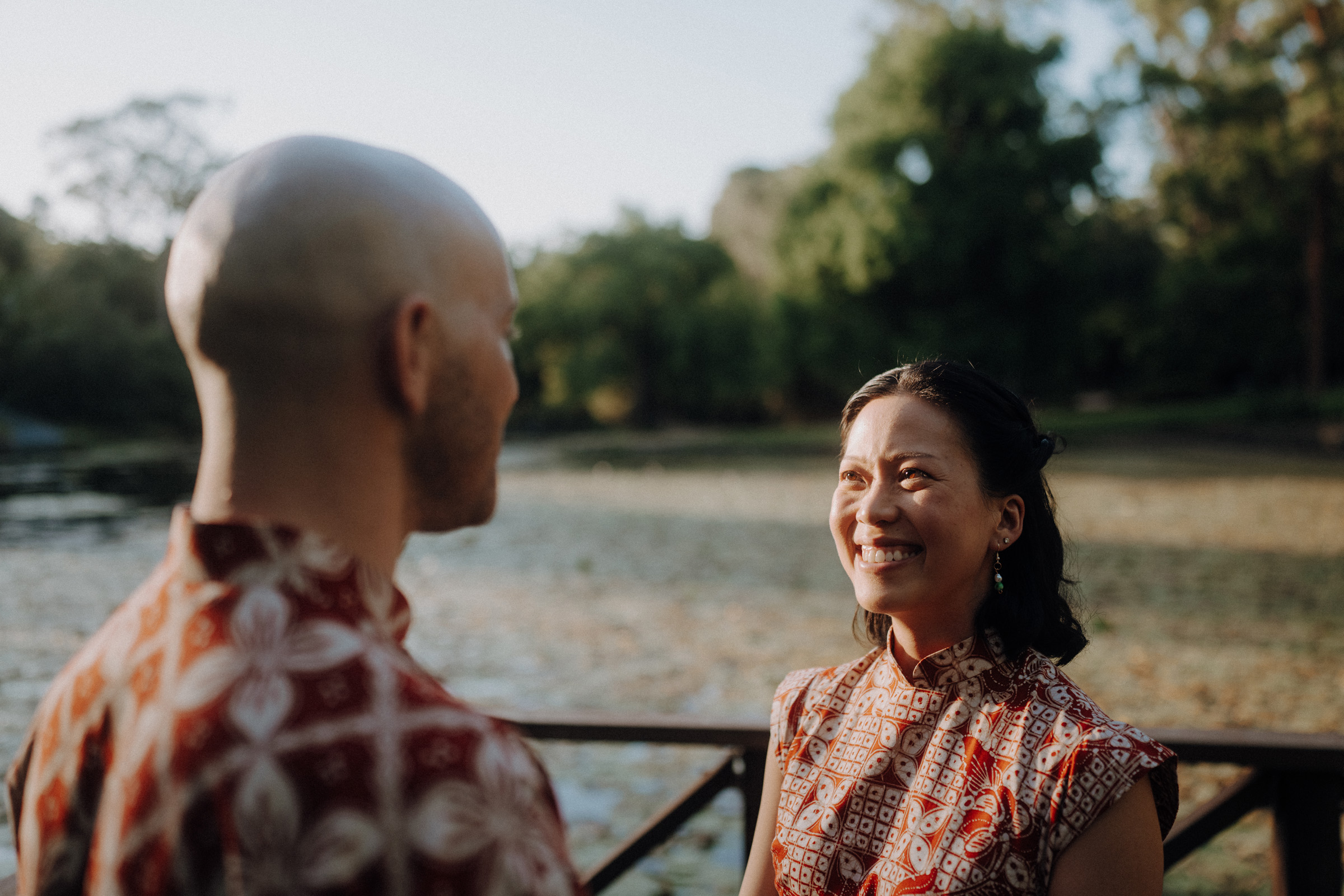 Two people wearing red patterned clothing stand on a wooden bridge by a body of water. The person on the right is smiling, with trees and greenery visible in the background.