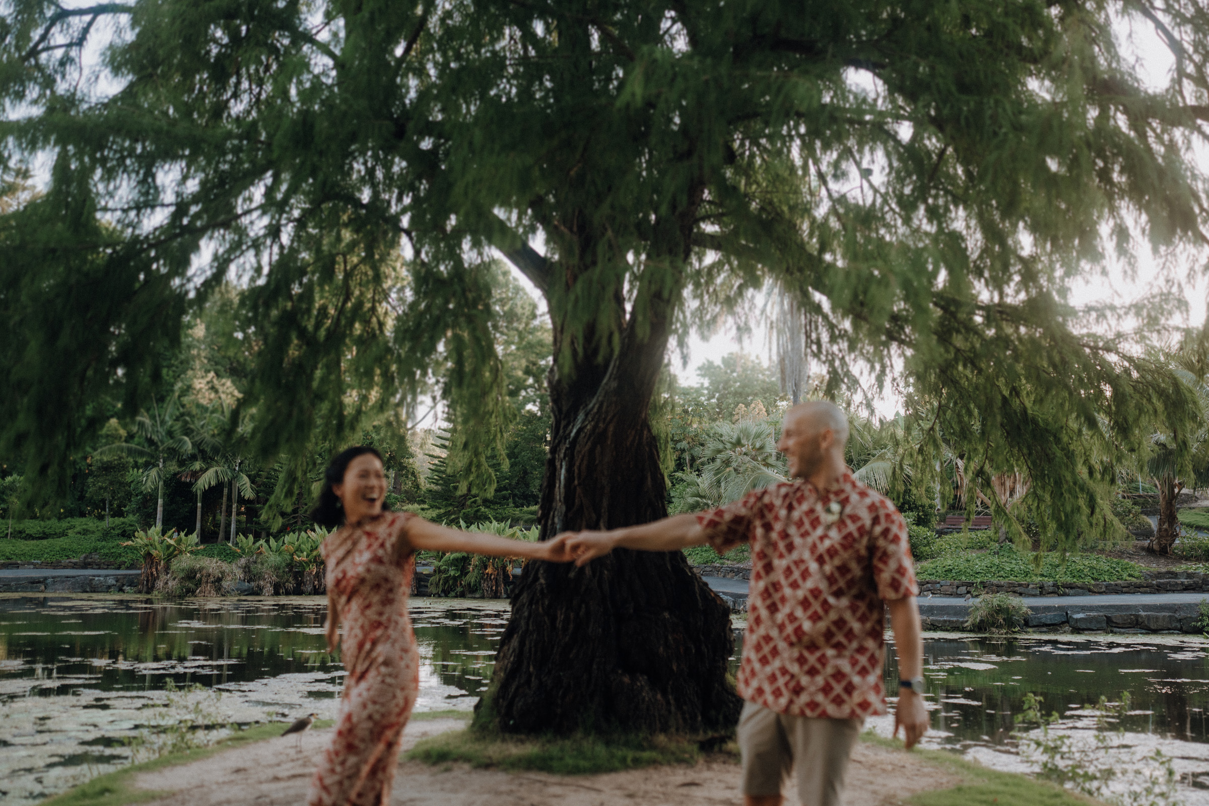 Two people wearing patterned outfits hold hands and walk in front of a large tree by a pond in a park setting.