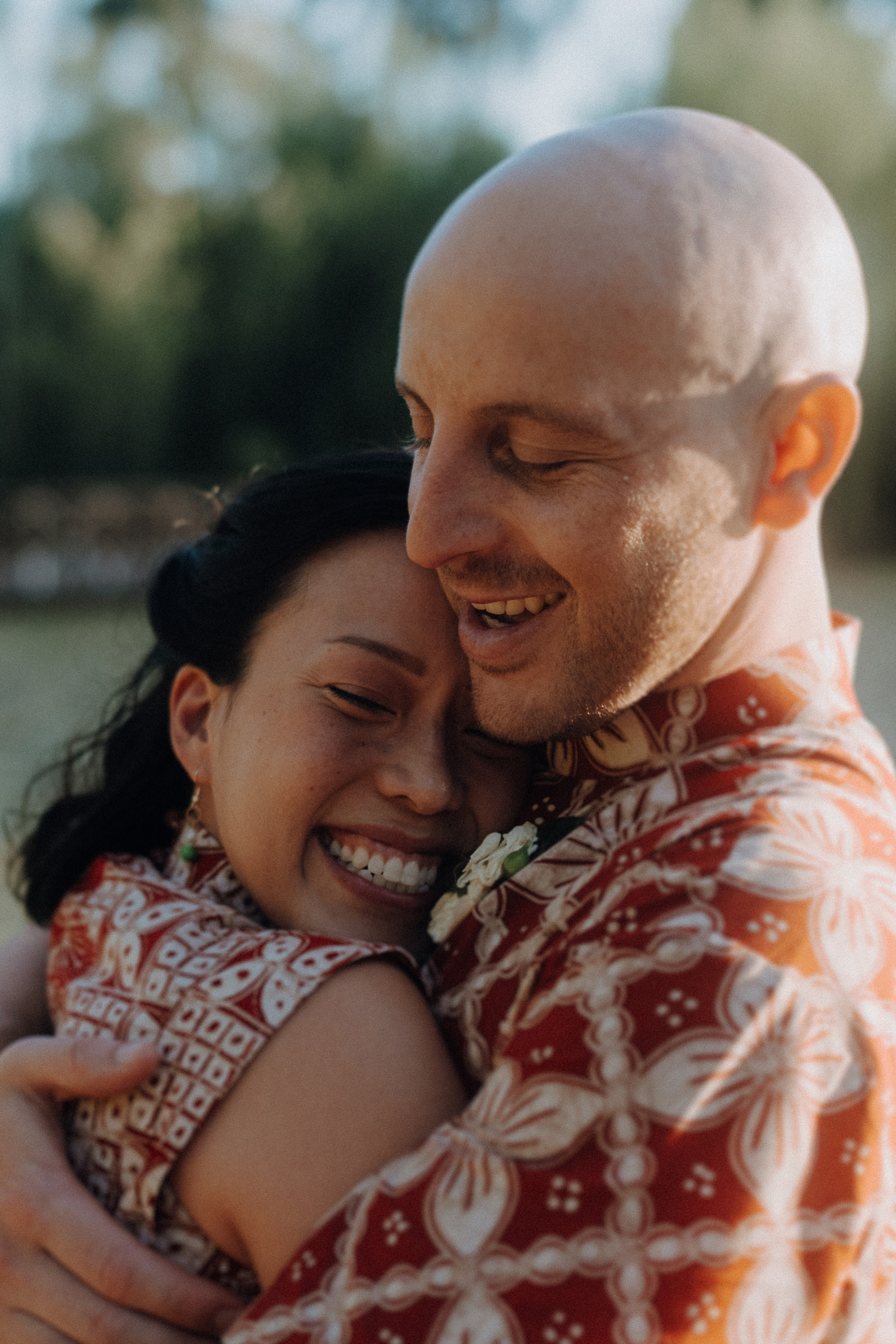 A couple embraces closely, both wearing patterned clothing. They are smiling and appear joyful, with a soft-focus green background.