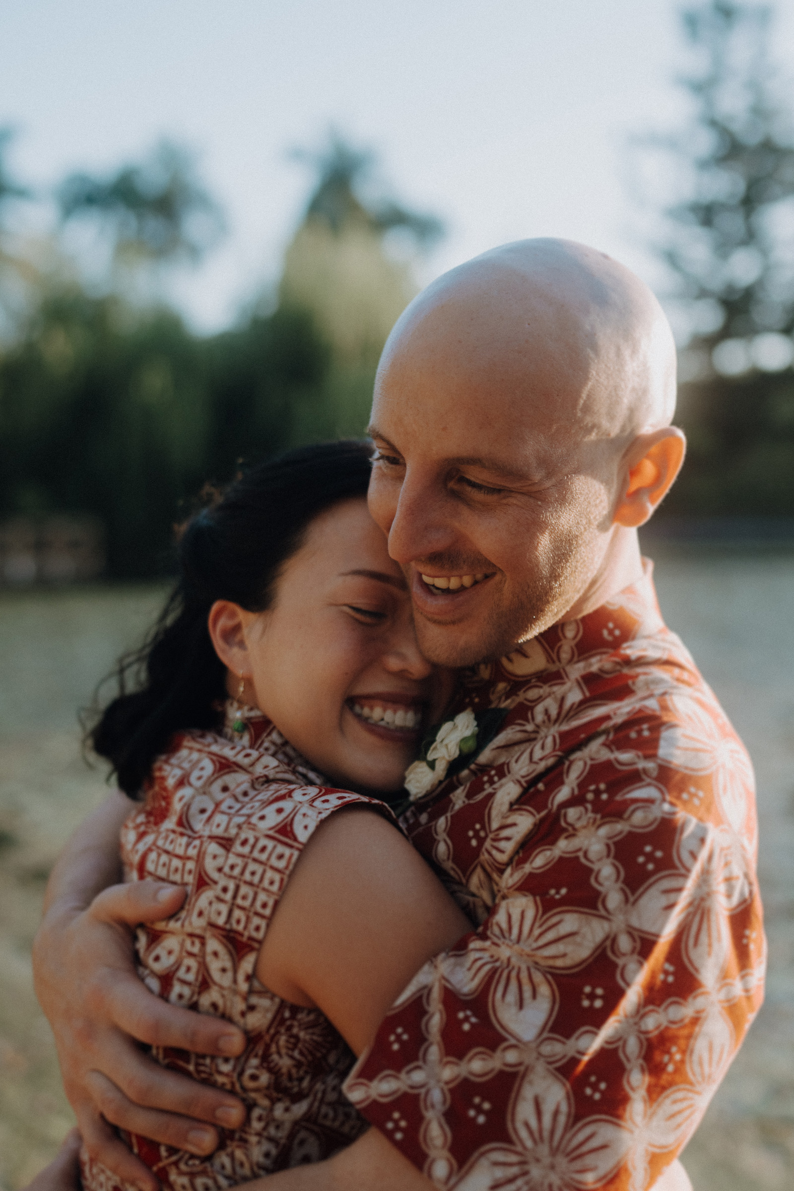 A couple in patterned shirts embrace and smile outdoors with trees in the background.