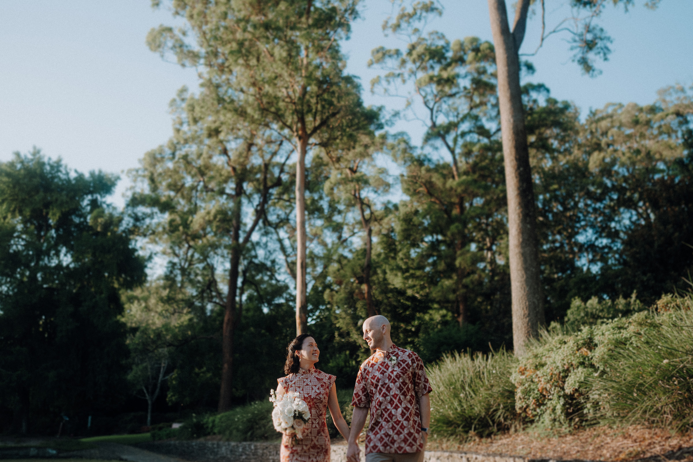 A couple in matching outfits holds hands, walking outdoors with trees in the background. The woman carries a bouquet.