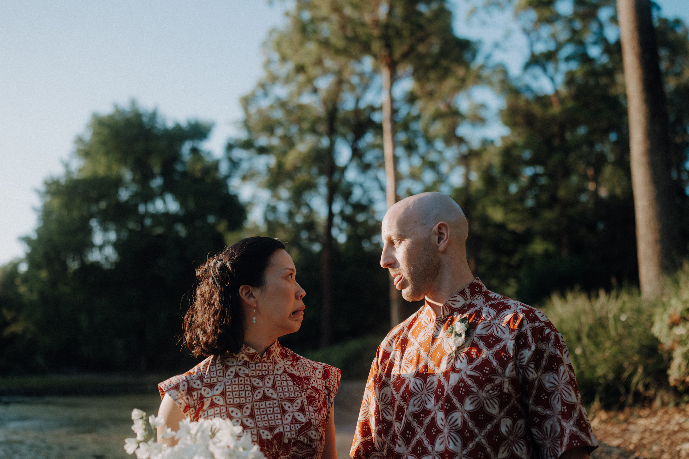 A couple in patterned shirts stand outside near trees, facing each other. The woman holds a bouquet of white flowers in her hand.