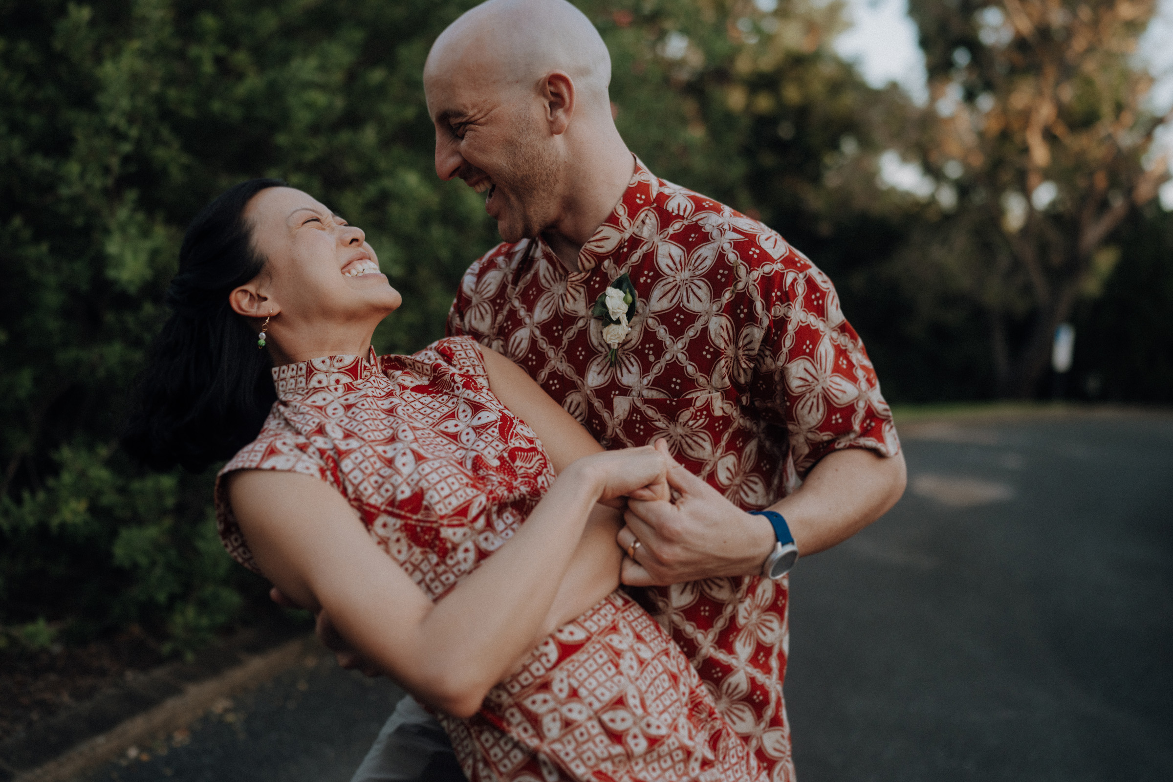 A couple in matching patterned outfits joyfully embrace on a road surrounded by trees.