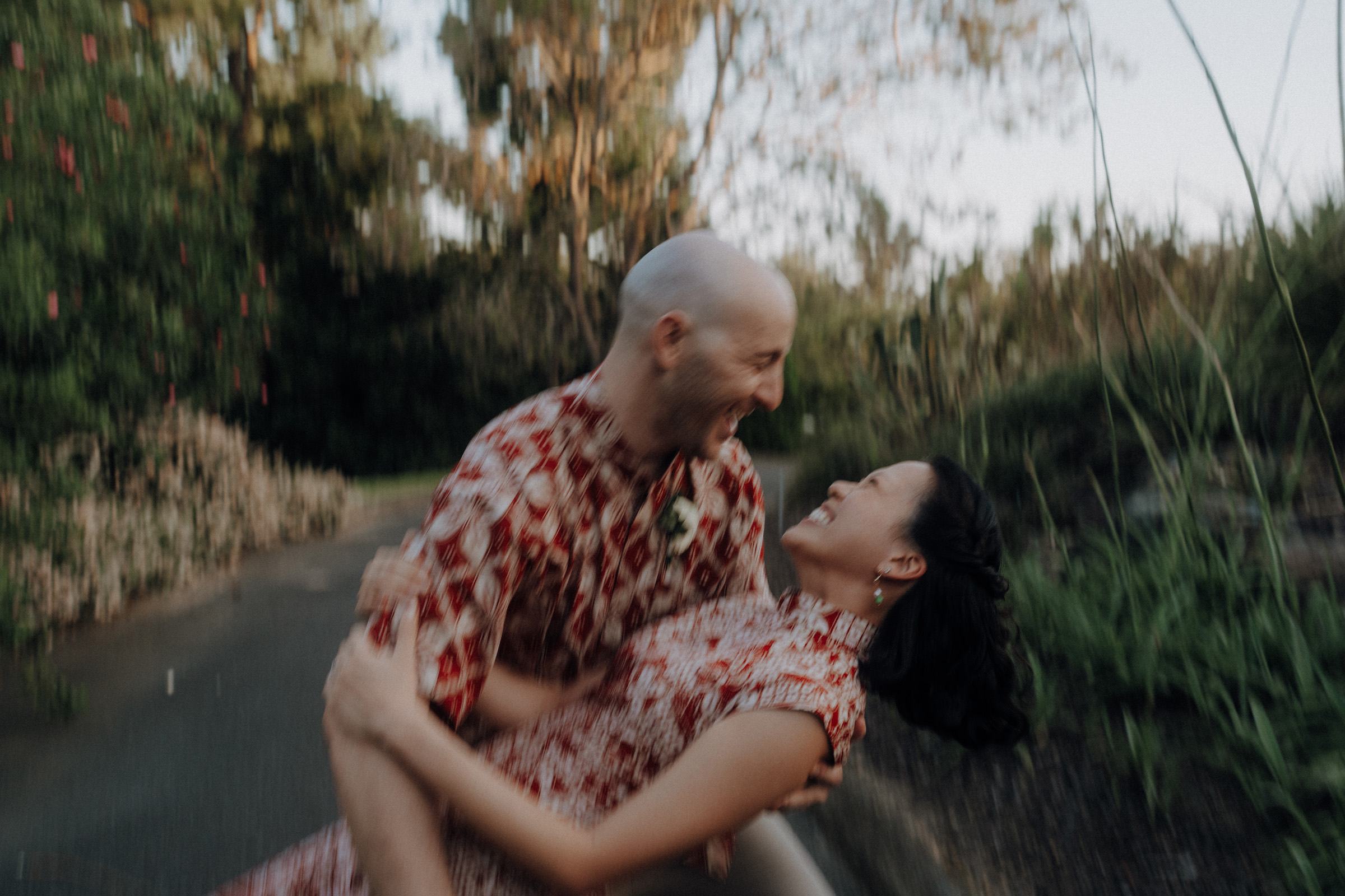 A couple in patterned outfits joyfully embracing on a pathway surrounded by greenery. The image is slightly blurred, conveying a sense of motion and happiness.