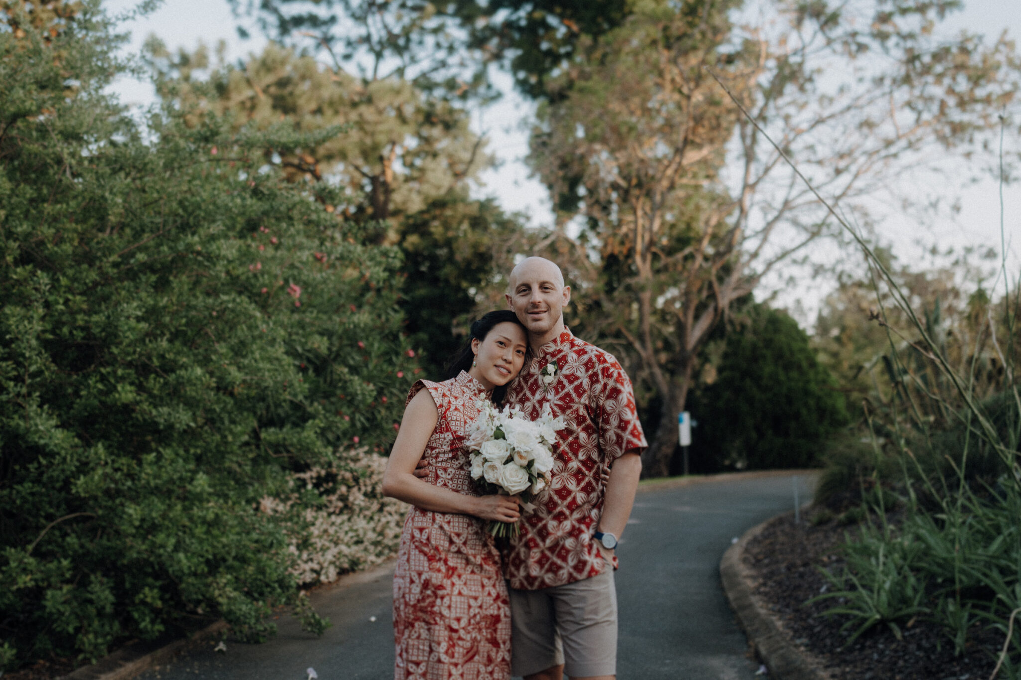 A couple stands on a path, the woman holding a bouquet of white flowers. Both wear patterned outfits. Greenery and trees are in the background.