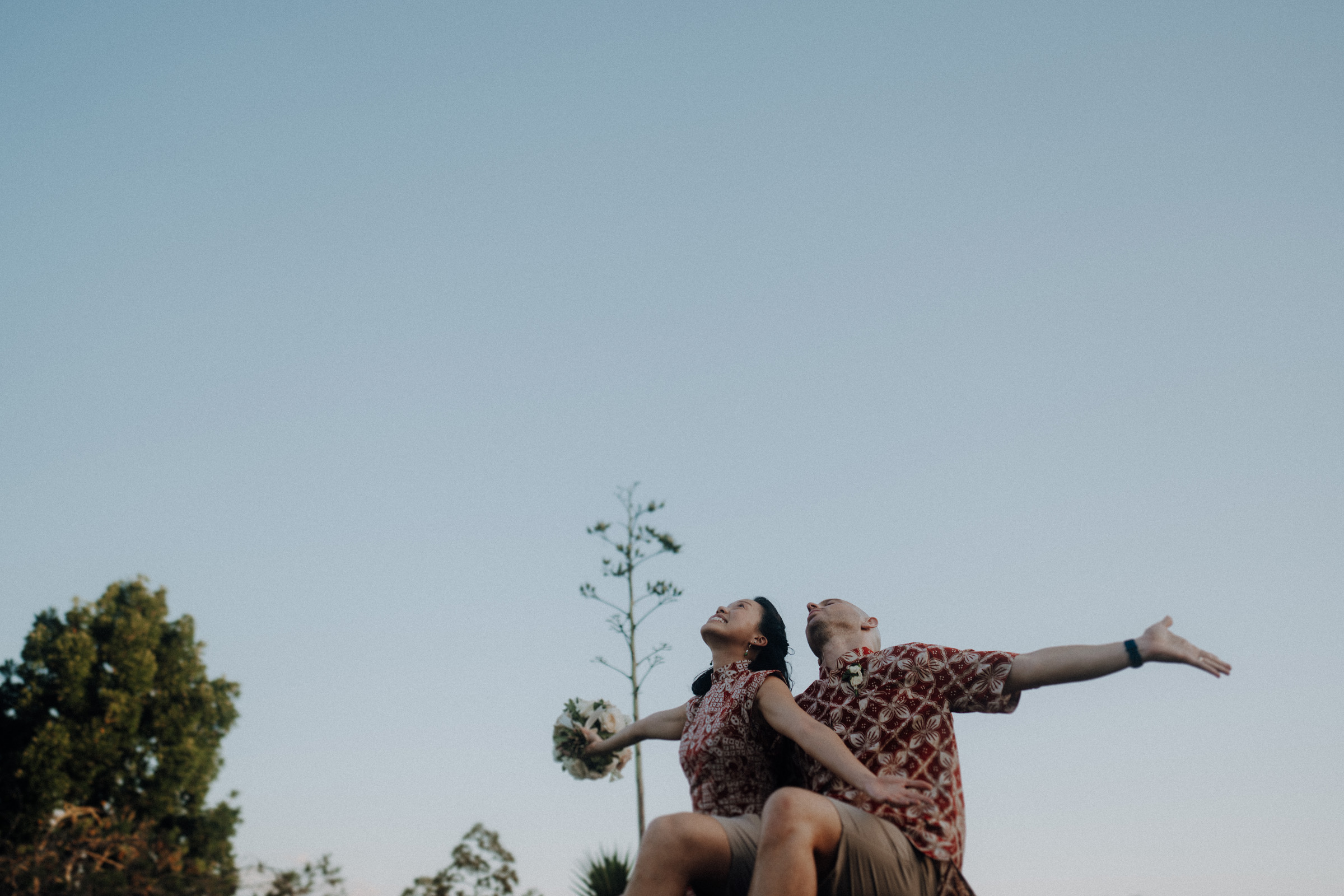 Two people in patterned shirts sit back-to-back on a grassy surface, arms outstretched, with a clear blue sky and trees in the background.