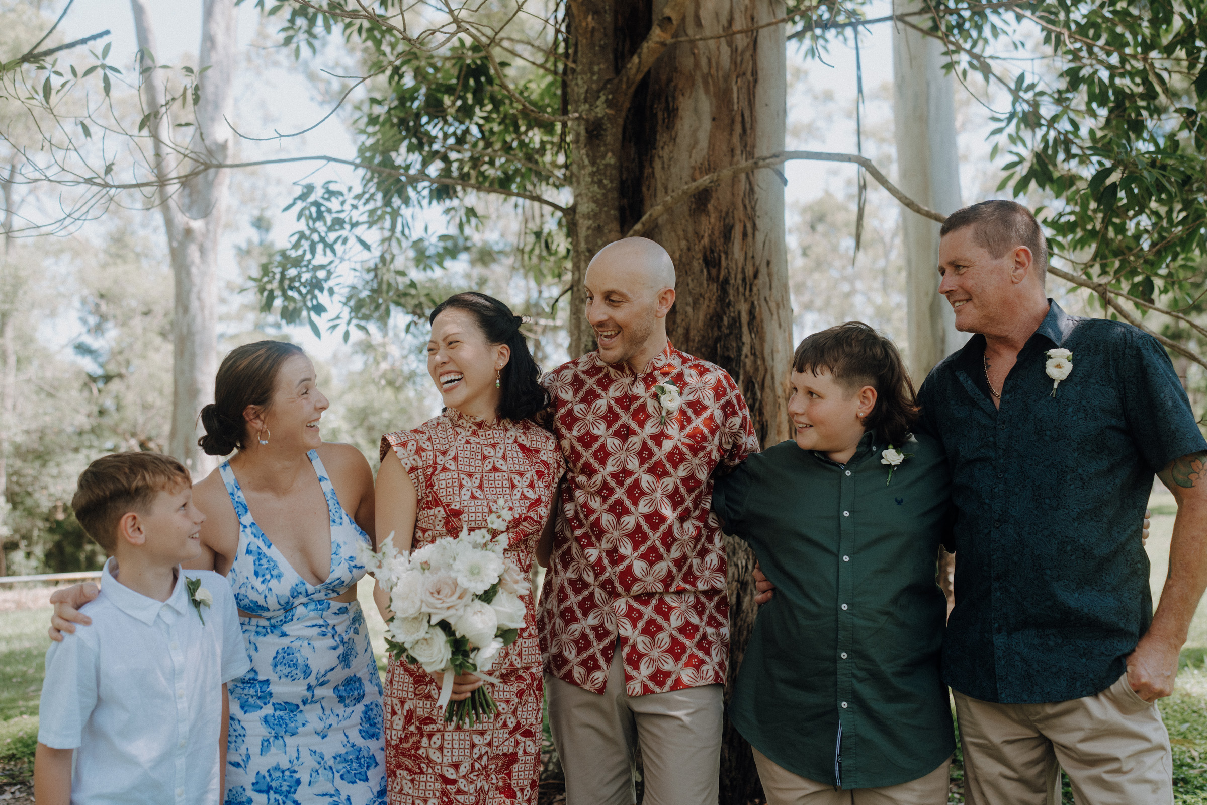 A group of people, including two children and four adults, stands outdoors near a tree, smiling and dressed in colorful attire. One adult holds a bouquet of flowers.