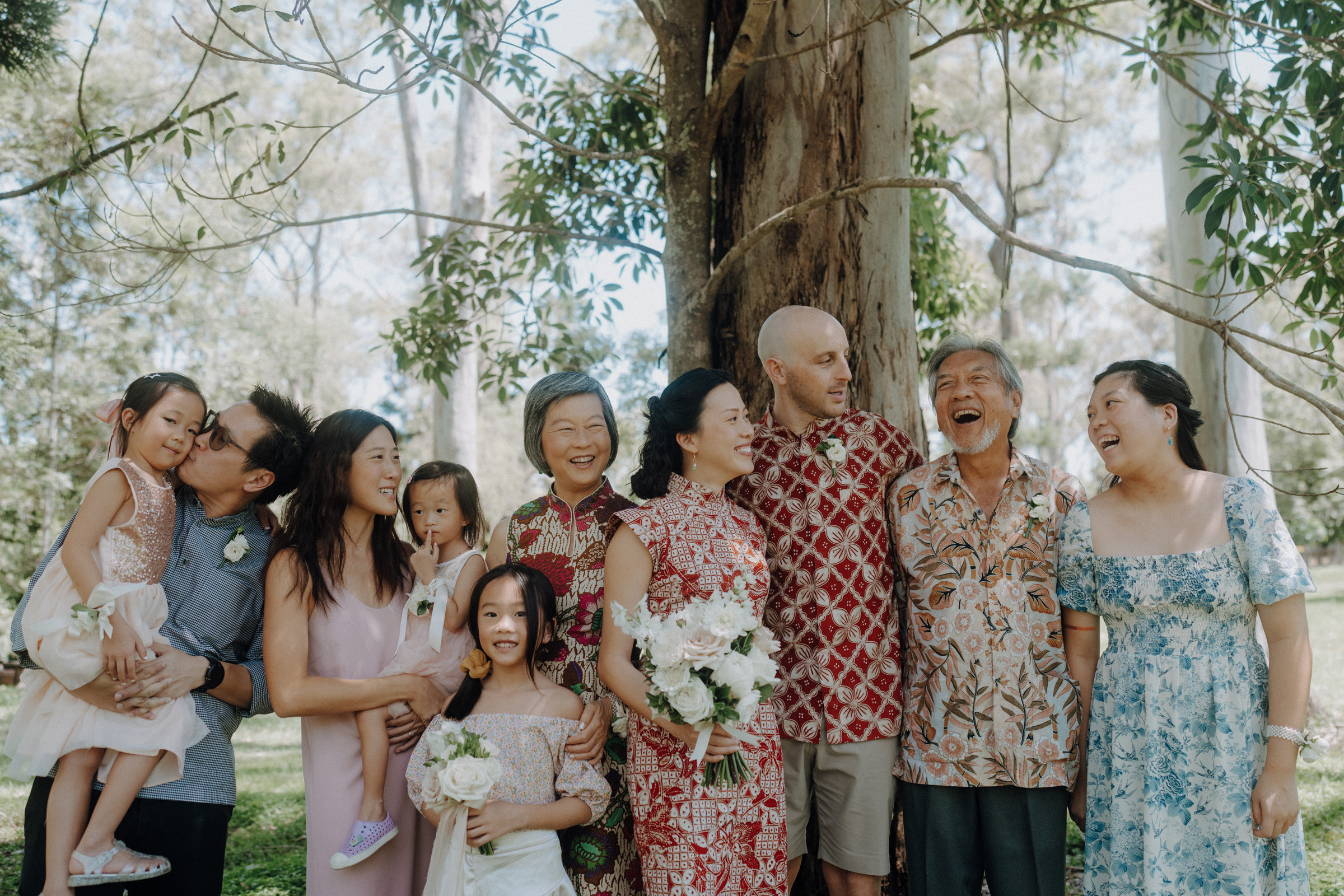 A group of people, including children and adults, stands outdoors in front of a tree, smiling and holding flowers. They are dressed in colorful, casual attire.