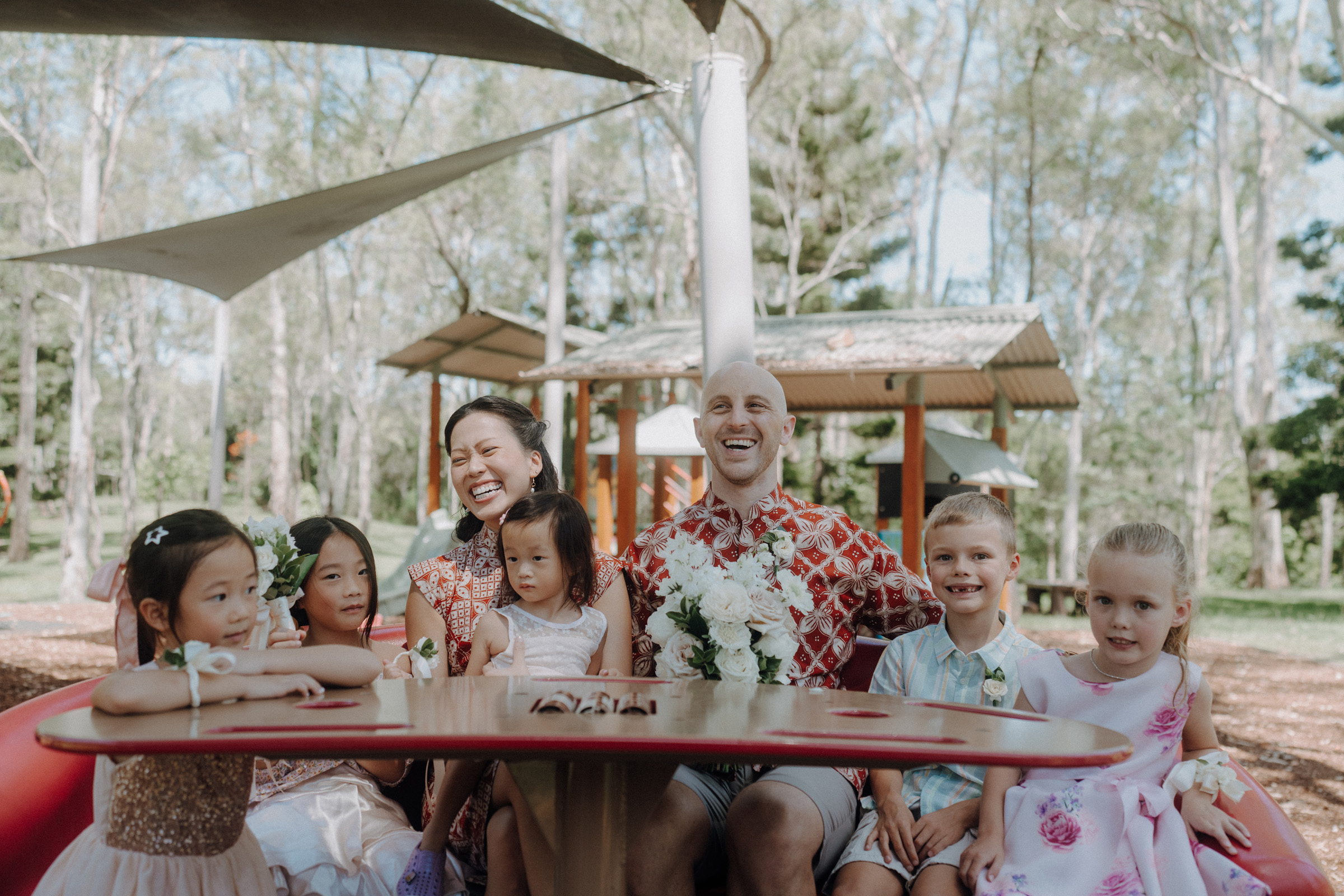 A group of adults and children sit at a round outdoor table under a shade structure, surrounded by trees, smiling and holding flowers.