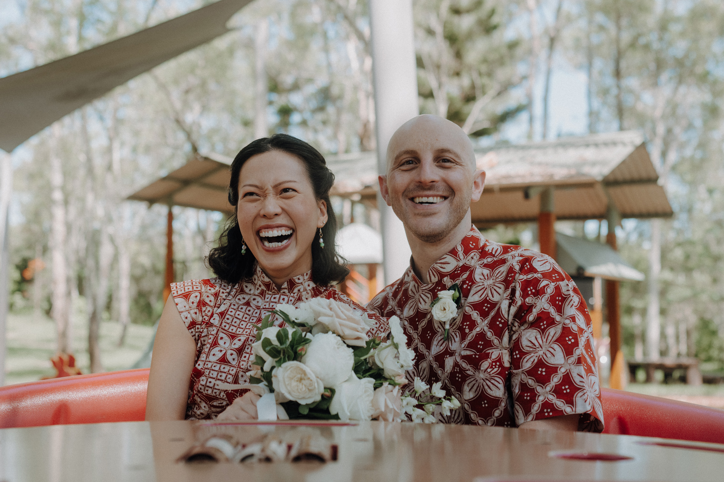 A couple in matching patterned outfits sits at a table outdoors, smiling and holding a bouquet of white flowers. Trees and a gazebo are in the background.