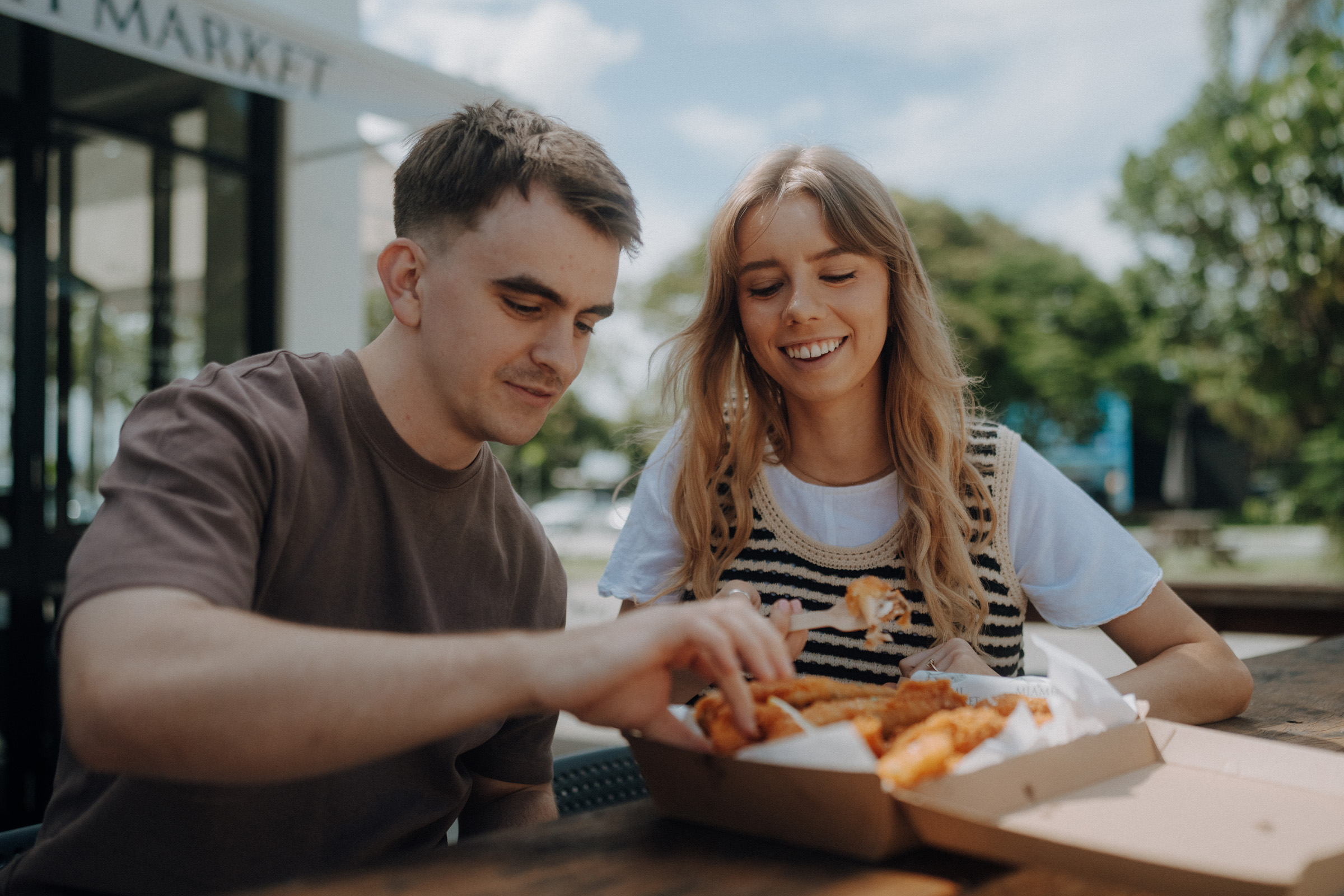 Two people are sitting outdoors at a table, sharing a box of food. The woman is smiling, and the man is picking up a piece of food. Trees and a building are visible in the background.