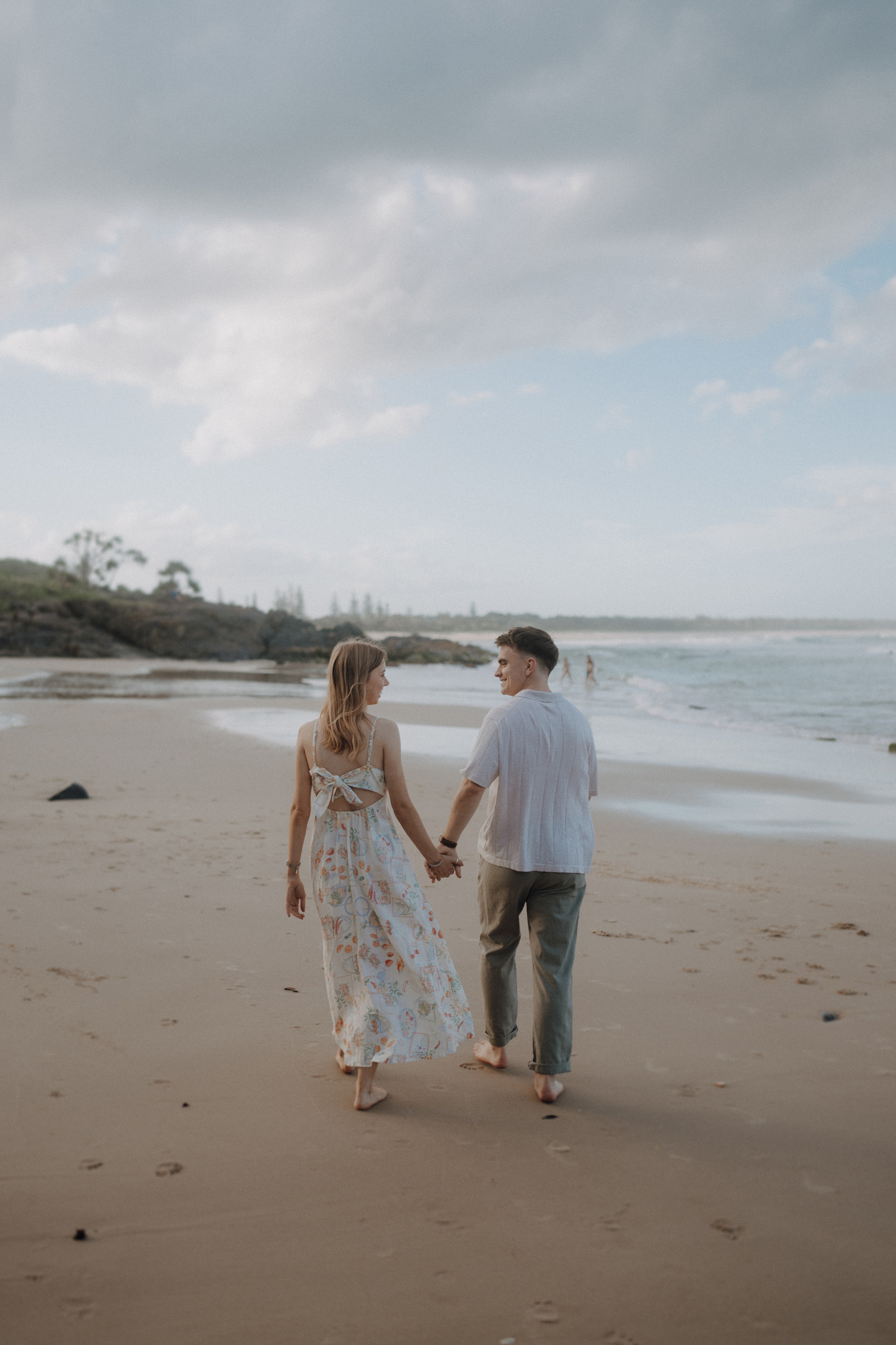 A couple holding hands walks along a beach with scattered clouds in the sky and waves in the background.