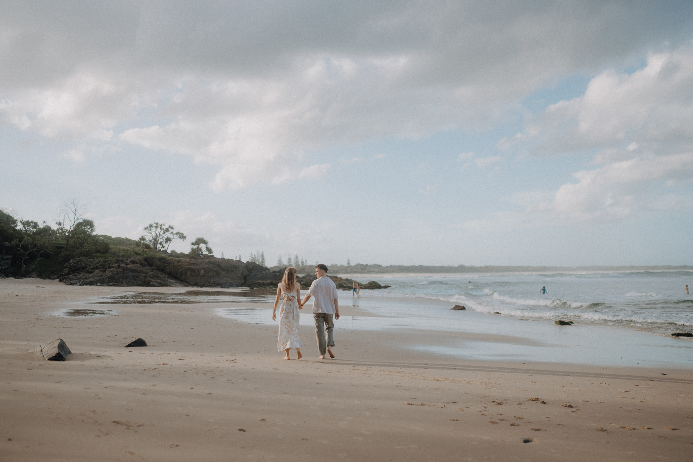 A couple walks along a sandy beach under a cloudy sky, with the ocean on their right and trees in the distance.