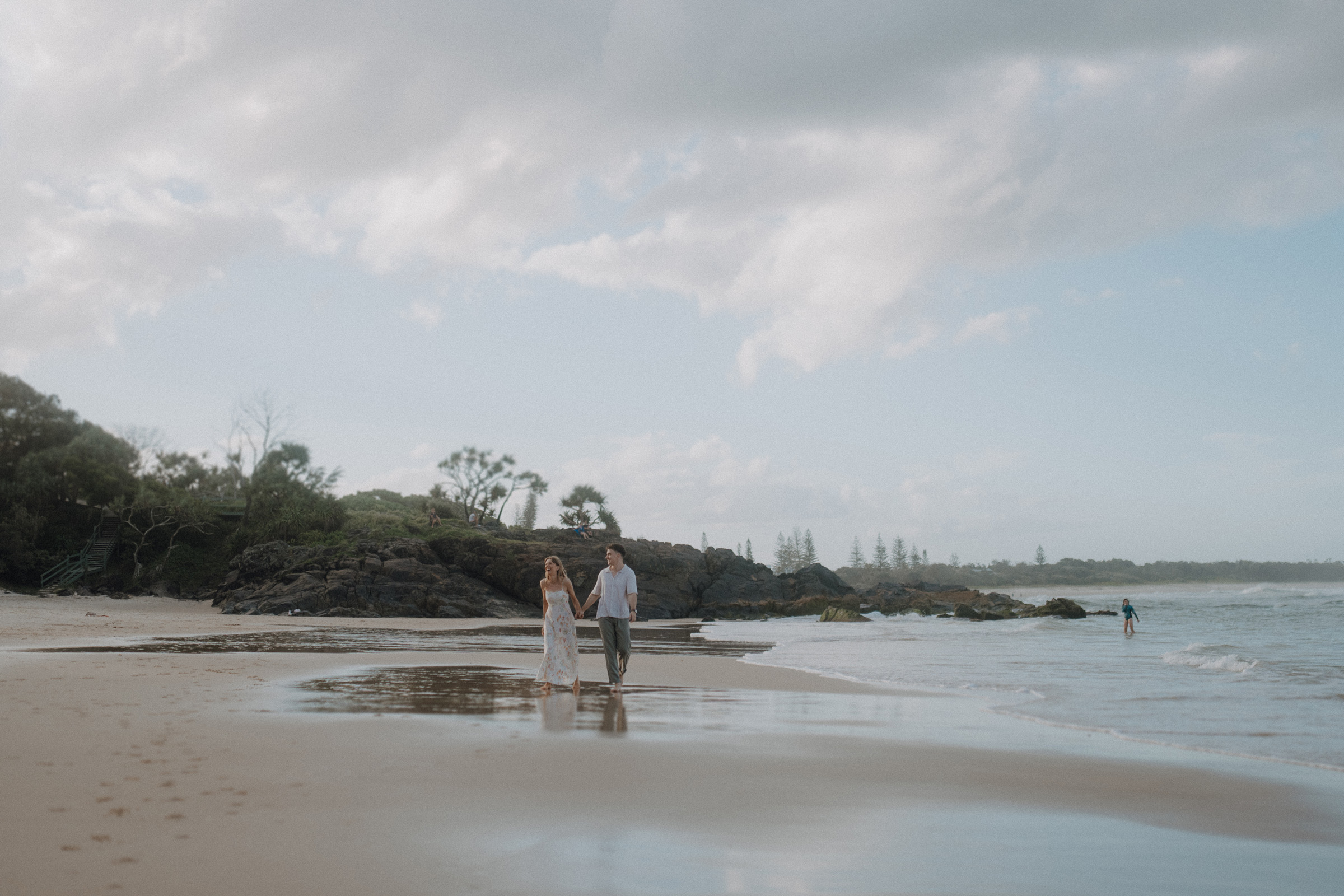 A couple walks along a sandy beach, holding hands, with waves nearby and distant trees under a partly cloudy sky.