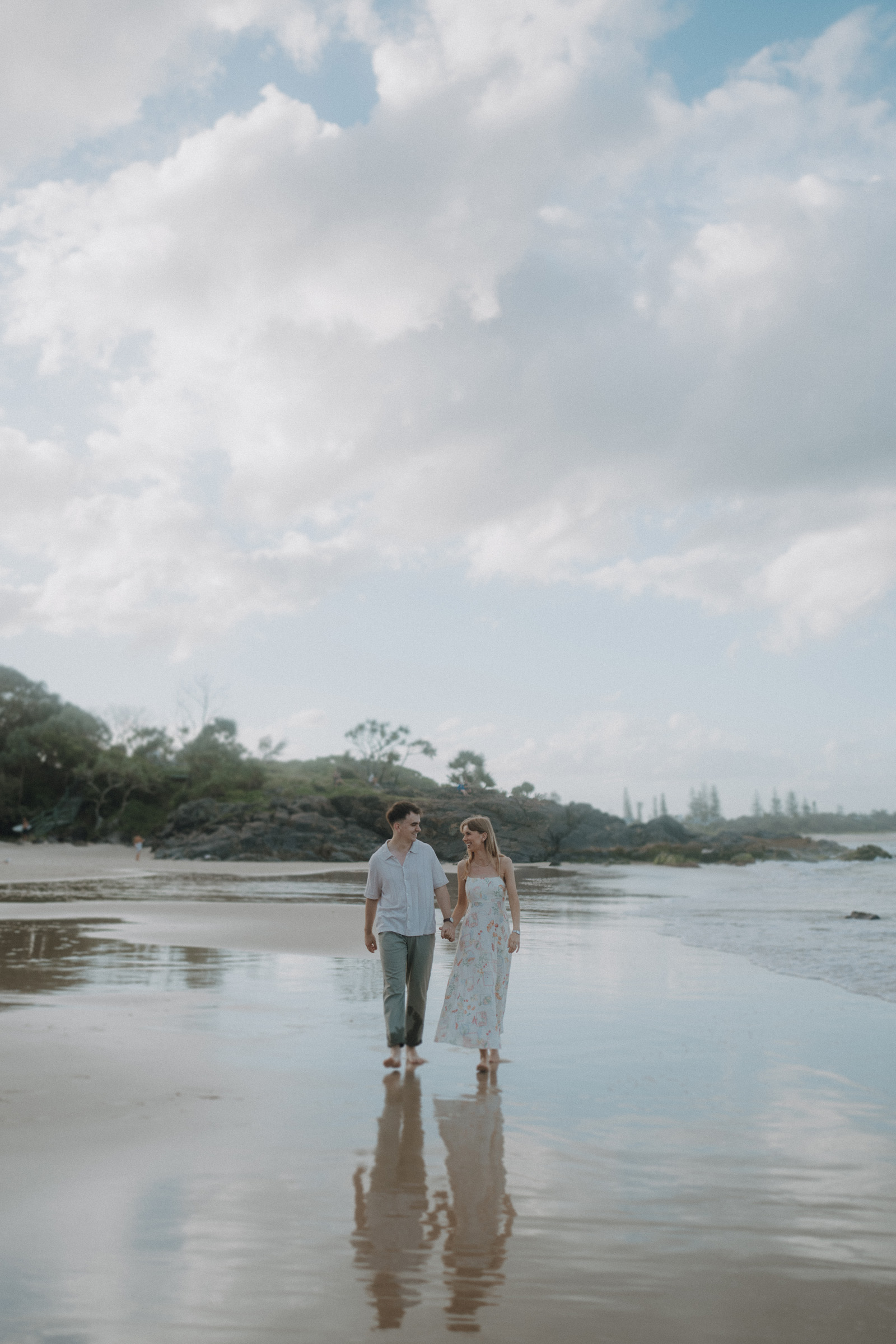 A couple holding hands walks along a sandy beach with clear skies and gentle waves.