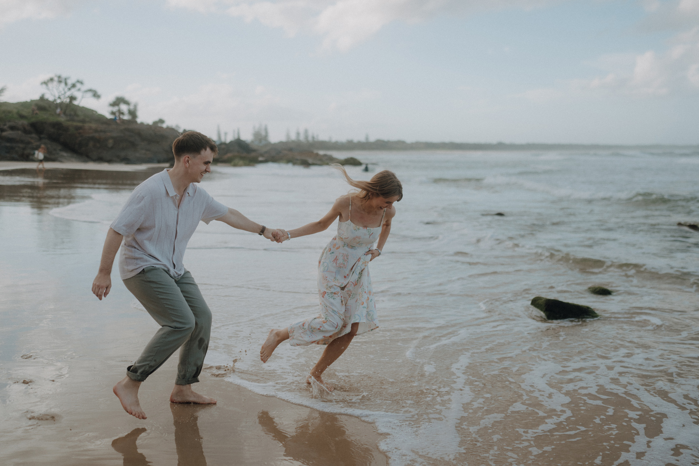 A couple holding hands and playfully running on a sandy beach near the ocean under a partly cloudy sky.