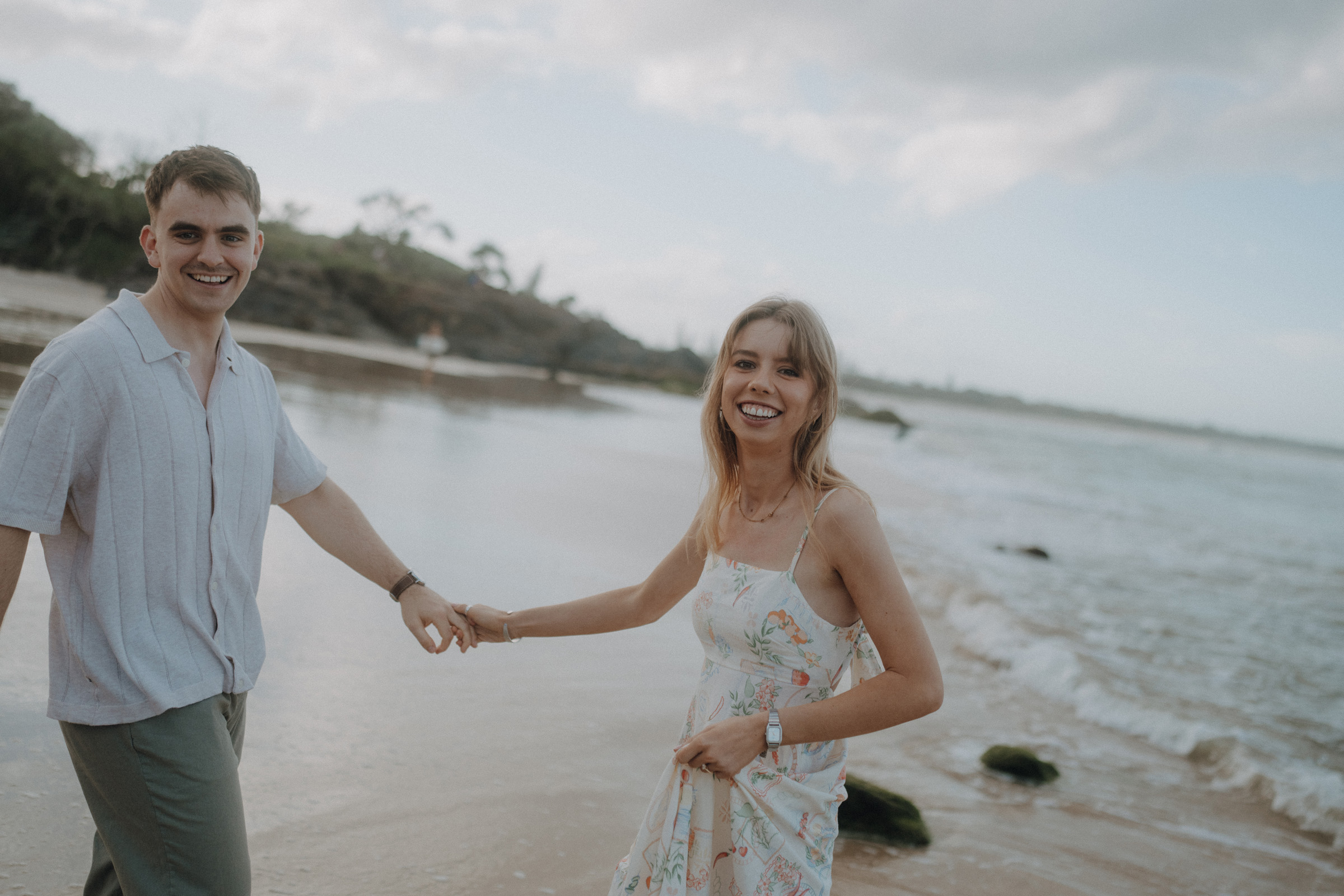 A man and a woman holding hands and smiling while walking on a beach, with the sea and a cloudy sky in the background.