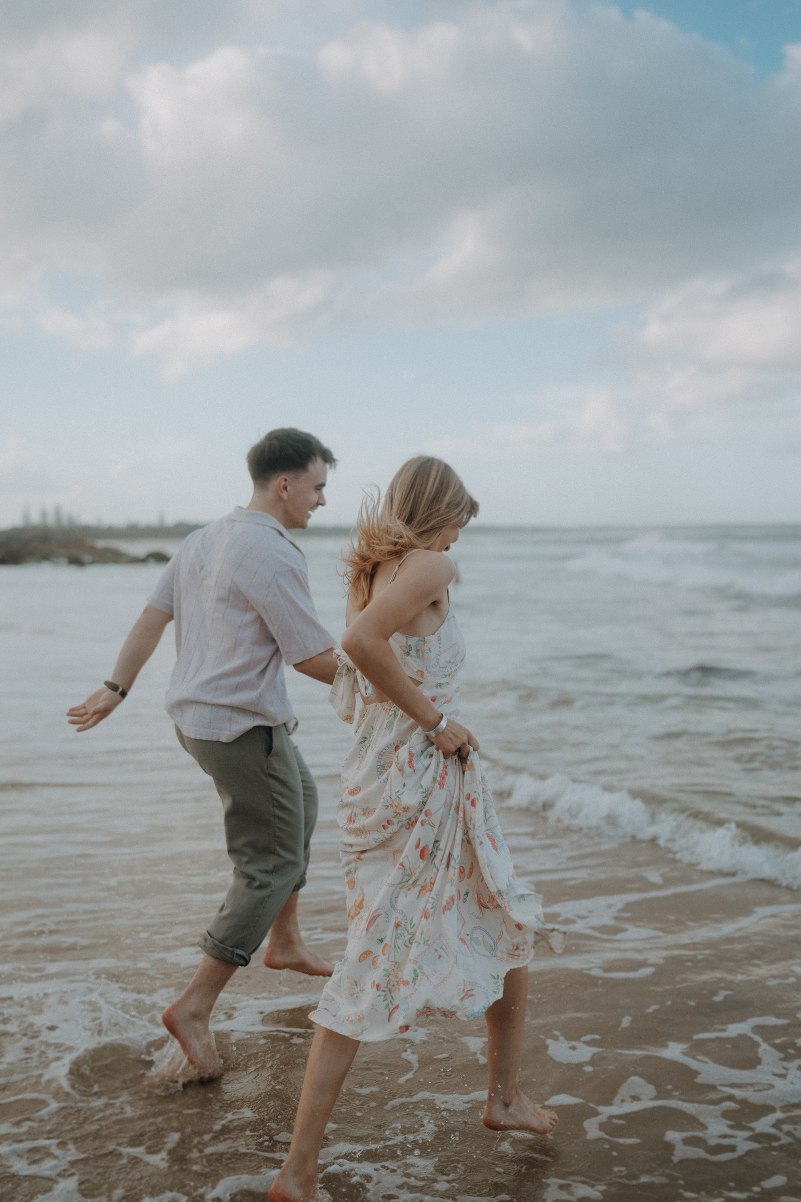 A man and woman walk barefoot along a sandy beach, with waves gently crashing nearby and a cloudy sky overhead.