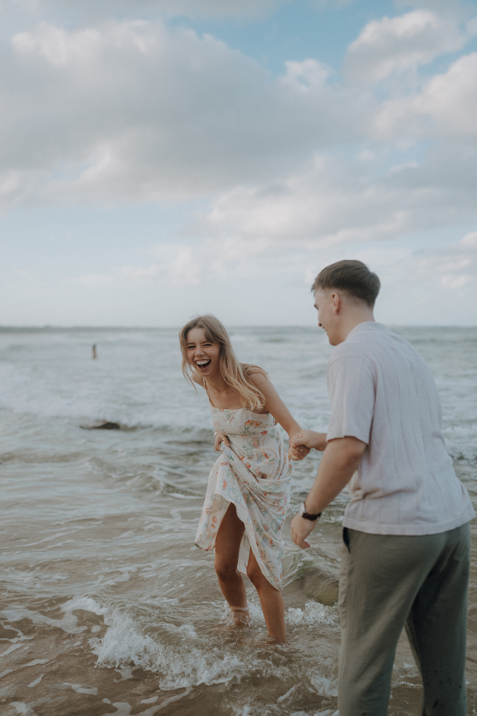 Two people standing at the beach, laughing and holding hands in shallow water. The sky is partly cloudy.