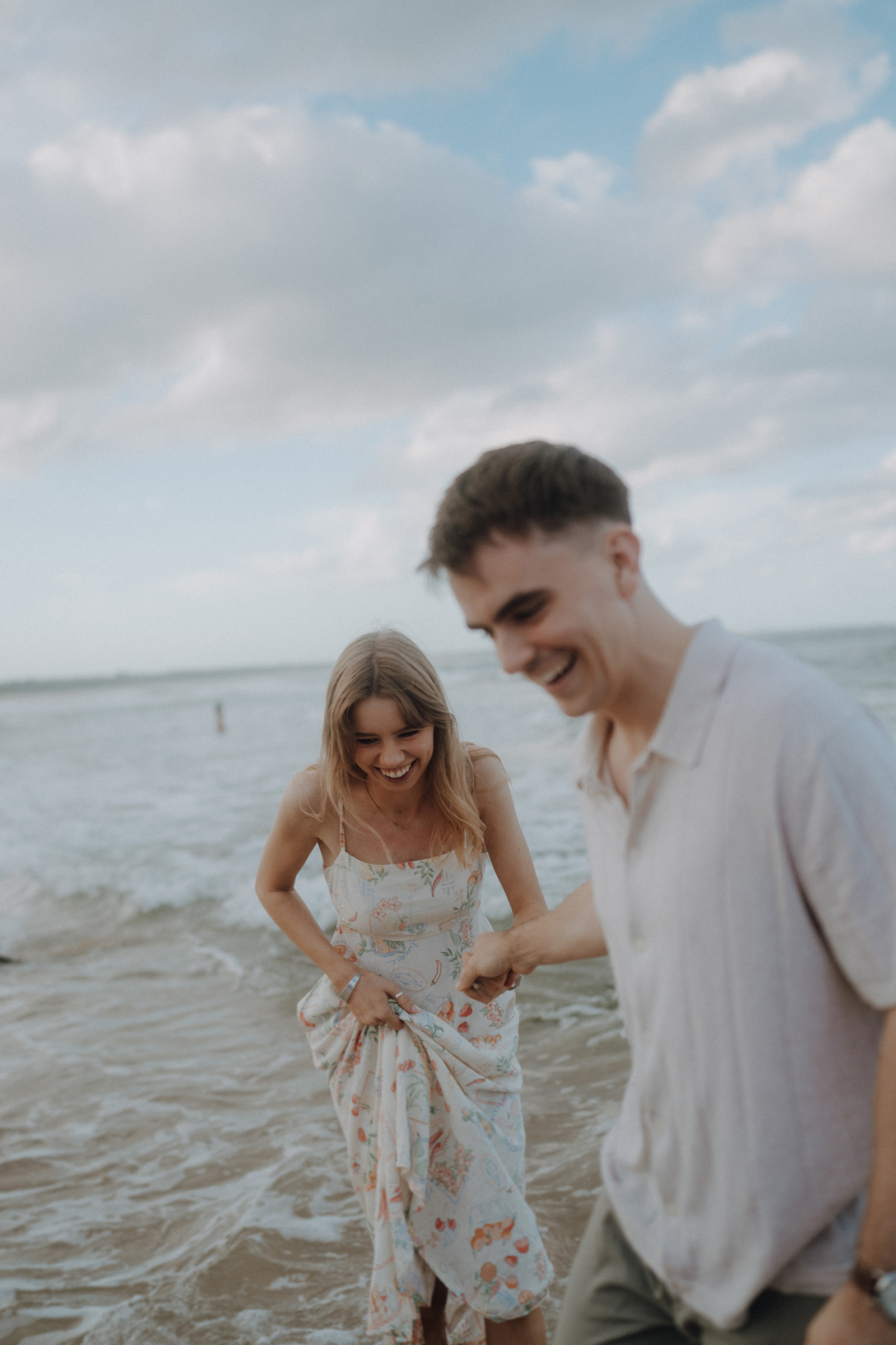 A woman in a floral dress and a man in a light shirt are laughing and walking on a beach with waves in the background.