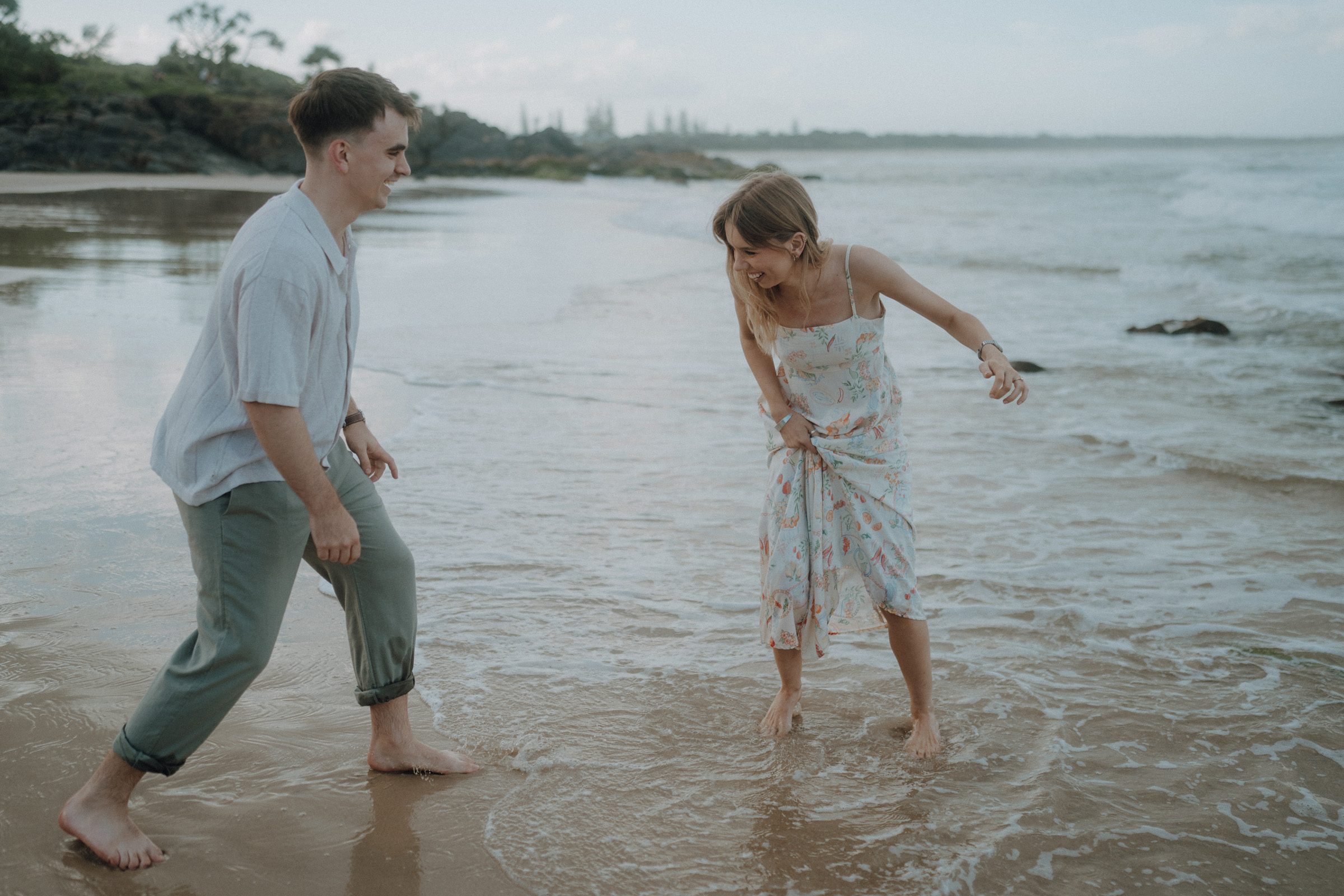 A man and woman stand barefoot on a beach, laughing as waves touch their feet. The sky is partly cloudy with a distant horizon of trees and rocks.