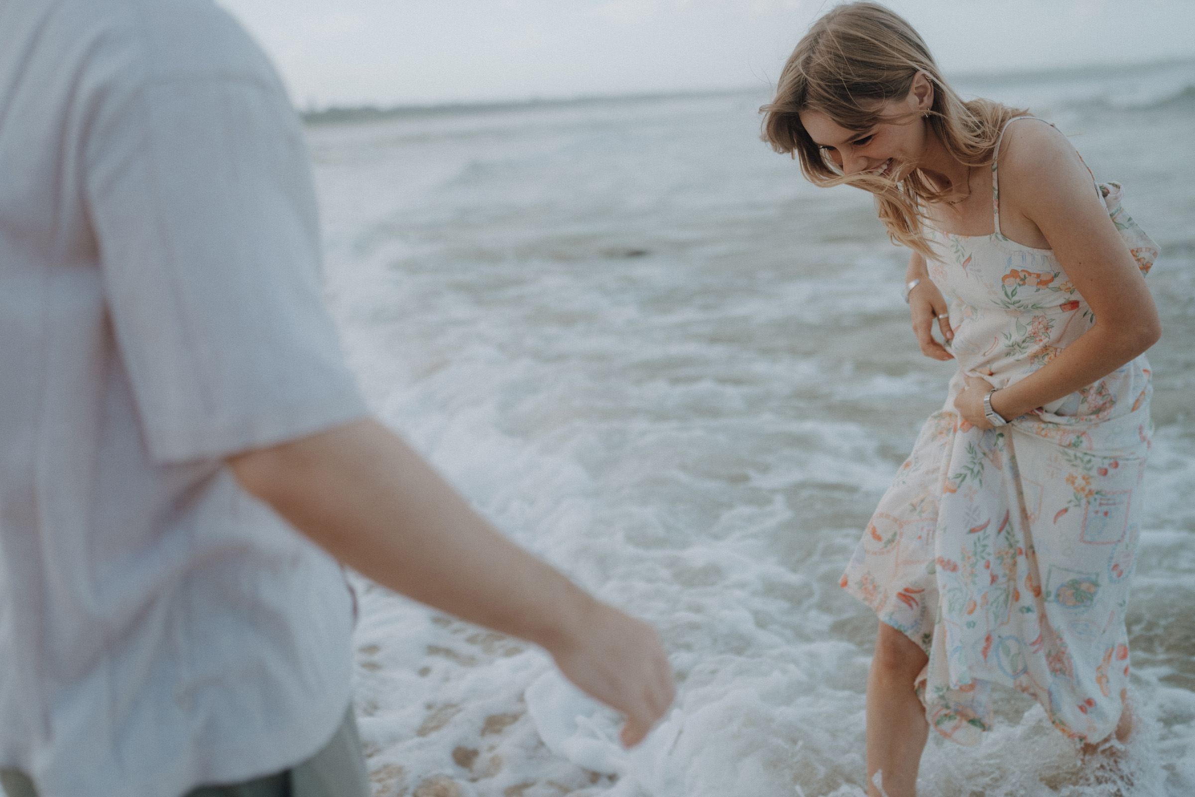 Two people stand at the edge of the ocean. The woman is wearing a floral dress and smiles while looking down at the water. The man is partially visible, extending his arm forward.