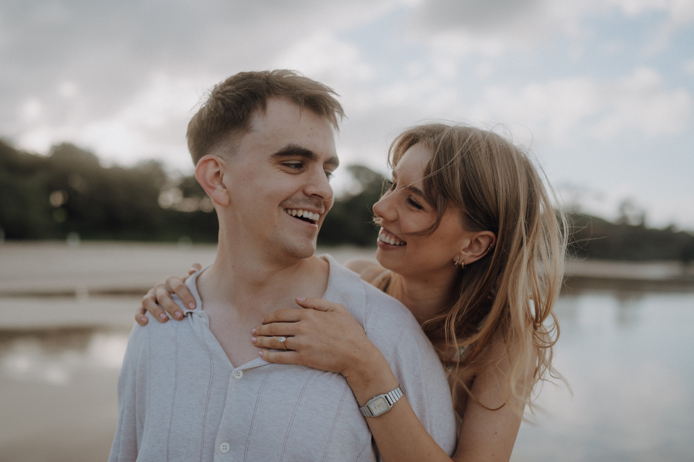 A woman embraces a smiling man from behind near a beach. Both appear happy, with blurred trees and water in the background.