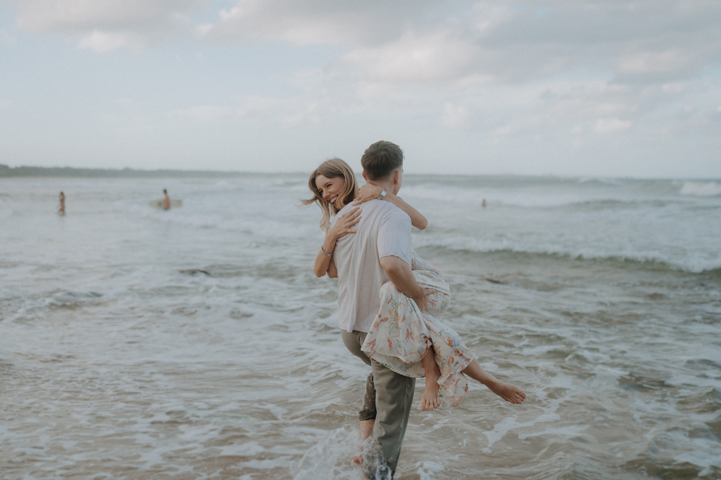 A man carries a woman through shallow ocean waves. She is holding onto his neck. The sky is cloudy, and a few people are in the background.