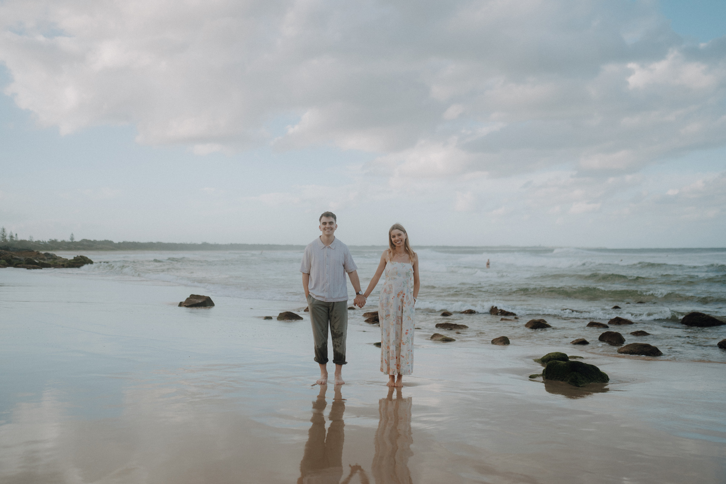 A couple holding hands stands on a rocky beach, with the ocean and a cloudy sky in the background.