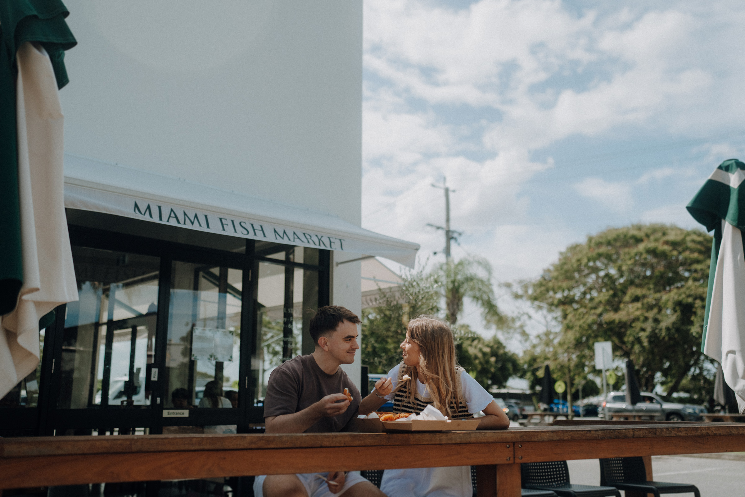 Two people sitting outdoors at a table, eating meals at the "Miami Fish Market." The scene includes a building with large windows and a backdrop of trees and a partly cloudy sky.