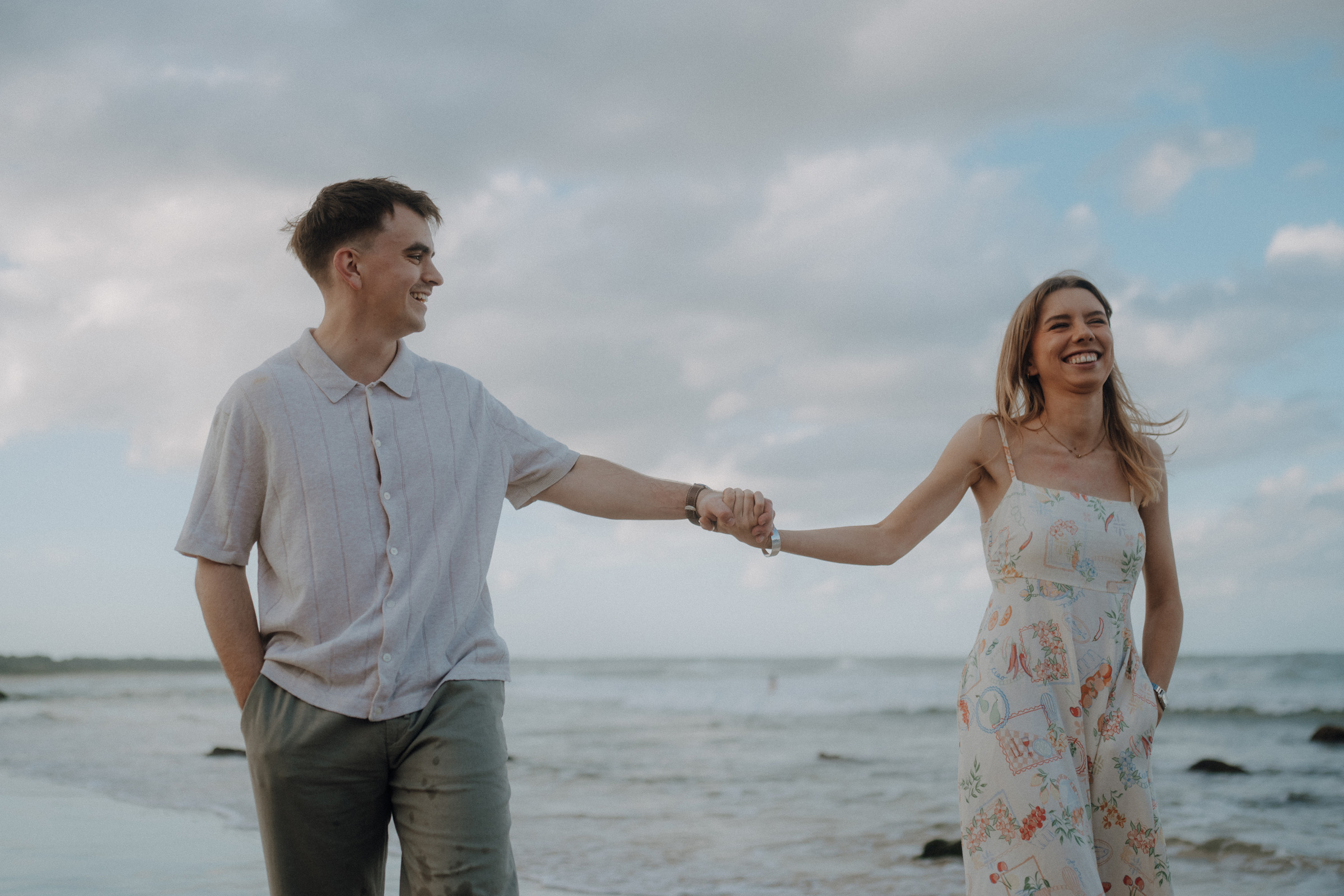 A couple holds hands and walks along a beach. The man wears a light shirt and pants, and the woman wears a patterned dress. The sky is partially cloudy.