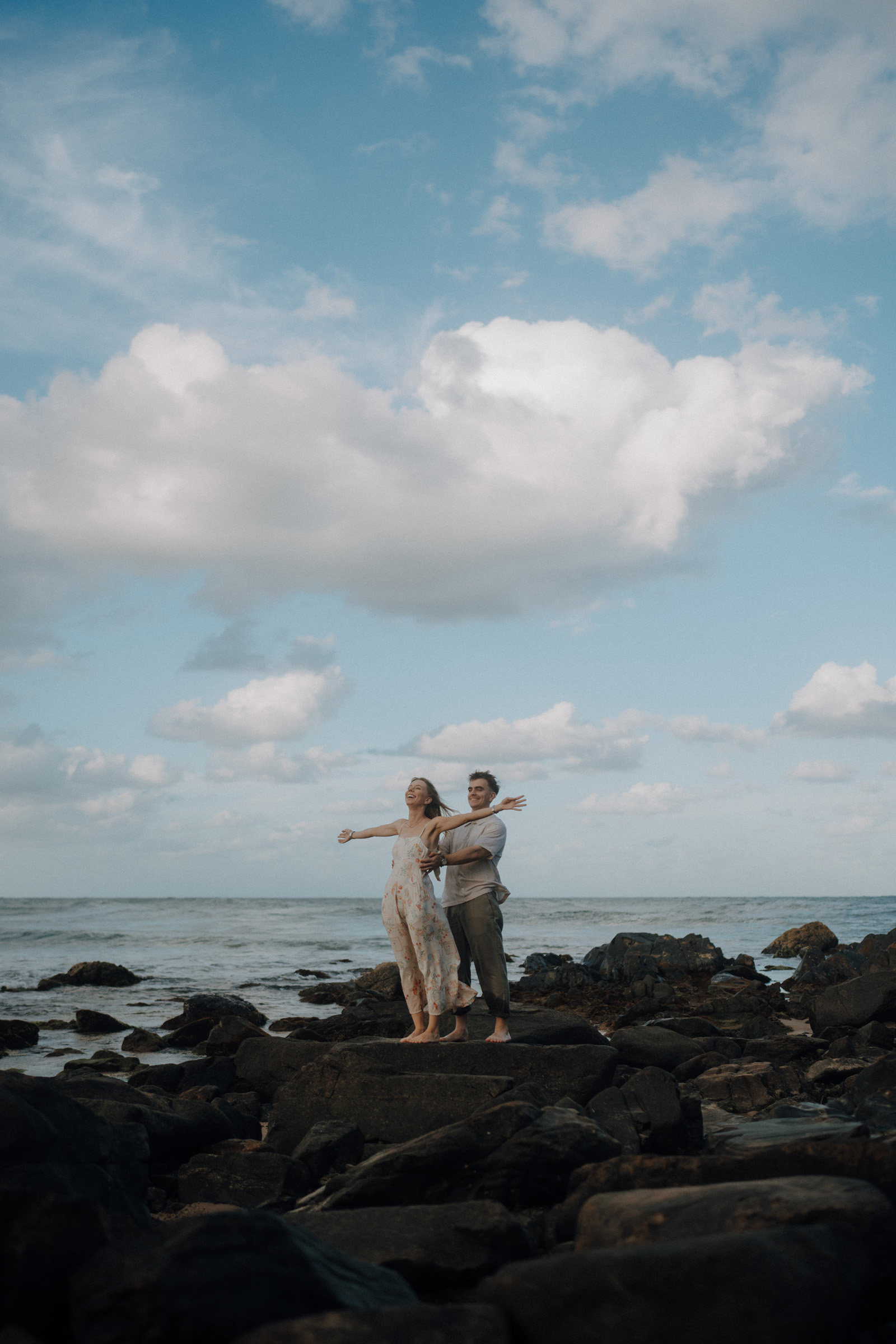 A couple stands on rocky shore with arms outstretched, facing the ocean under a partly cloudy sky.