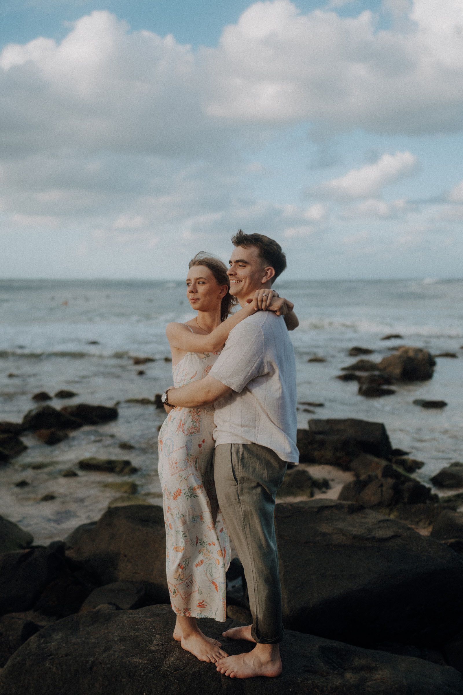 A couple embraces on rocky shorelines, with the ocean and a cloudy sky in the background.