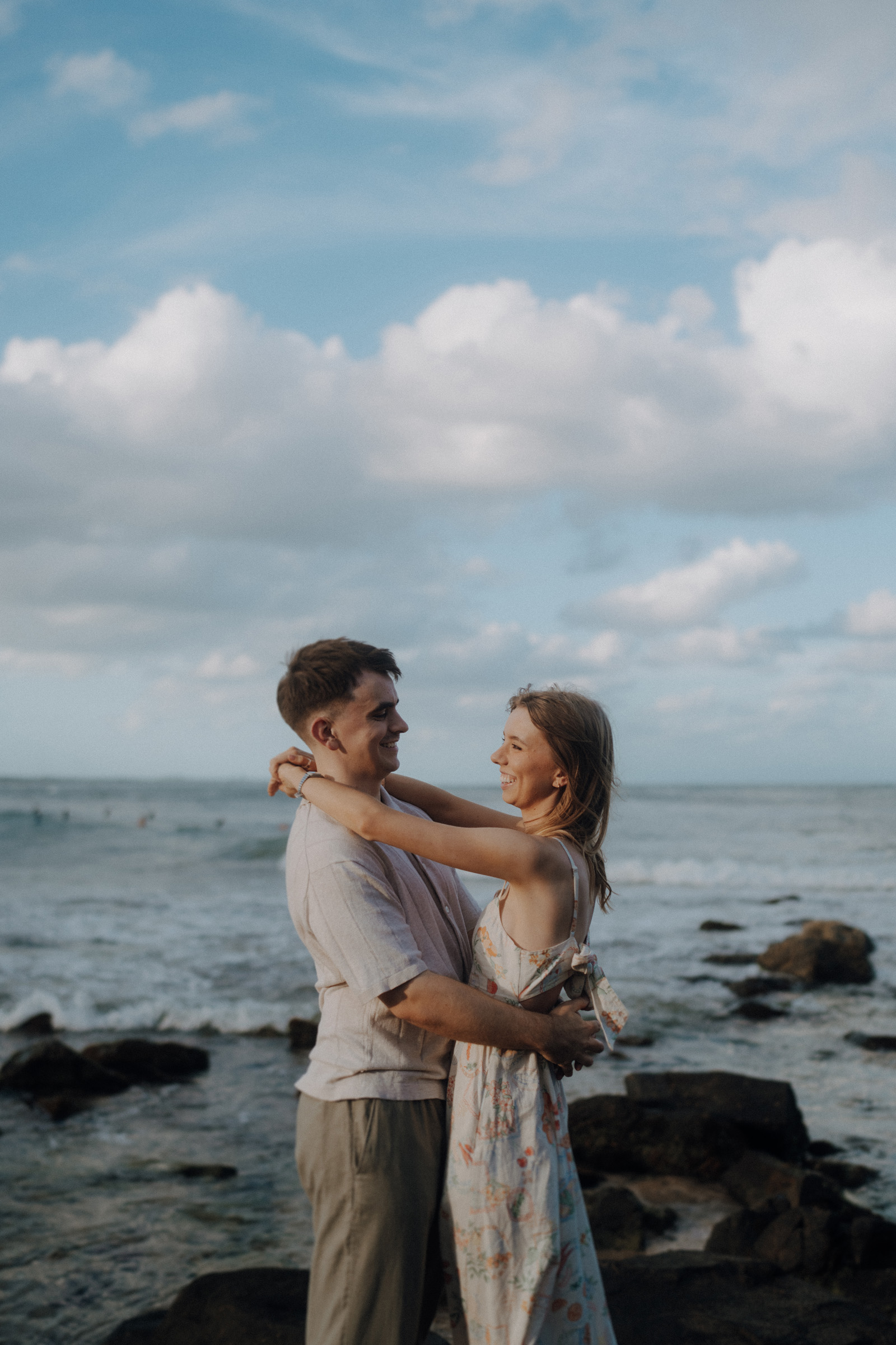 A couple embraces on a rocky shore with the ocean and a partly cloudy sky in the background.