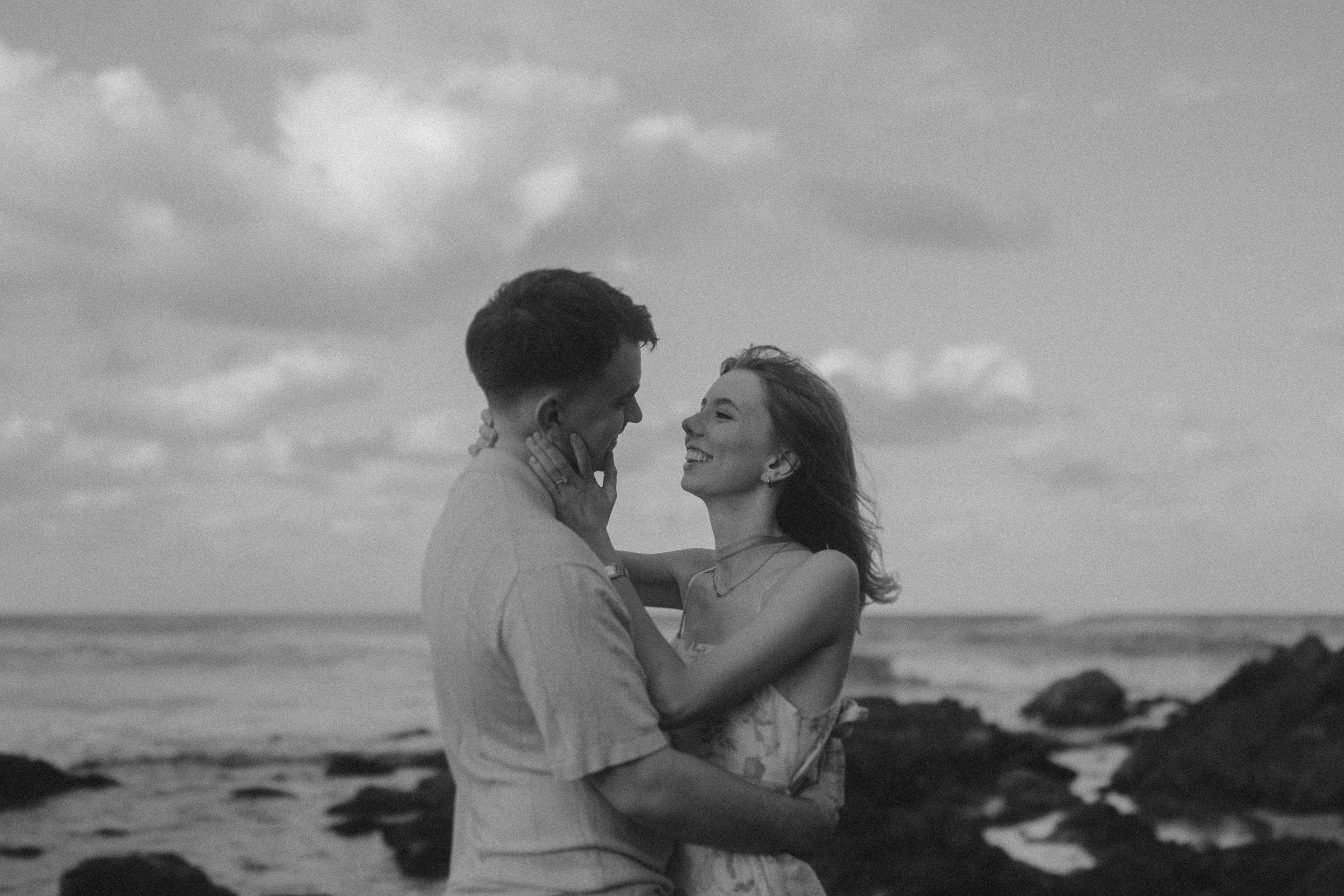 A couple embraces and smiles at each other on a rocky beach with the ocean and cloudy sky in the background. The image is in black and white.