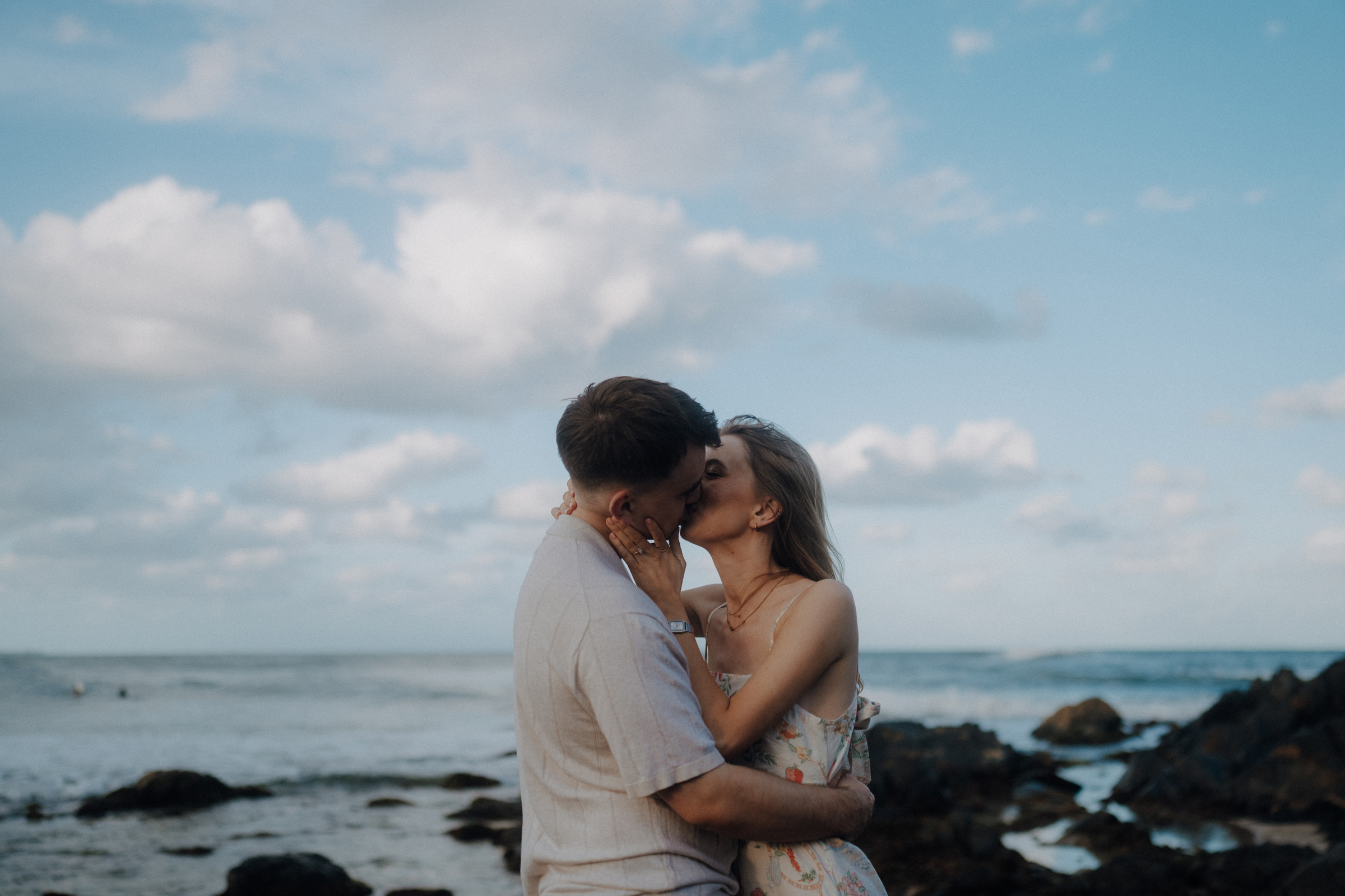 A couple embraces and kisses on a rocky beach under a cloudy sky.