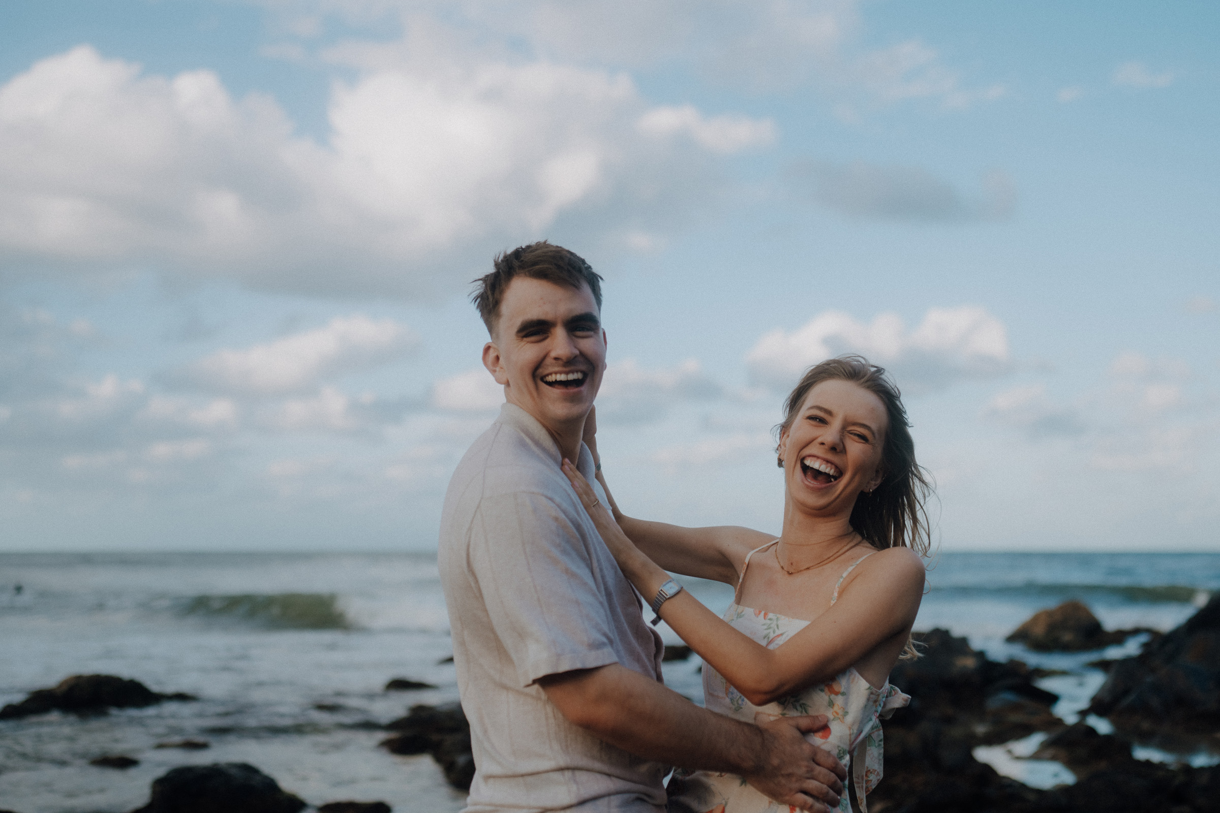 A couple laughing joyfully near the ocean, with clouds and waves in the background.