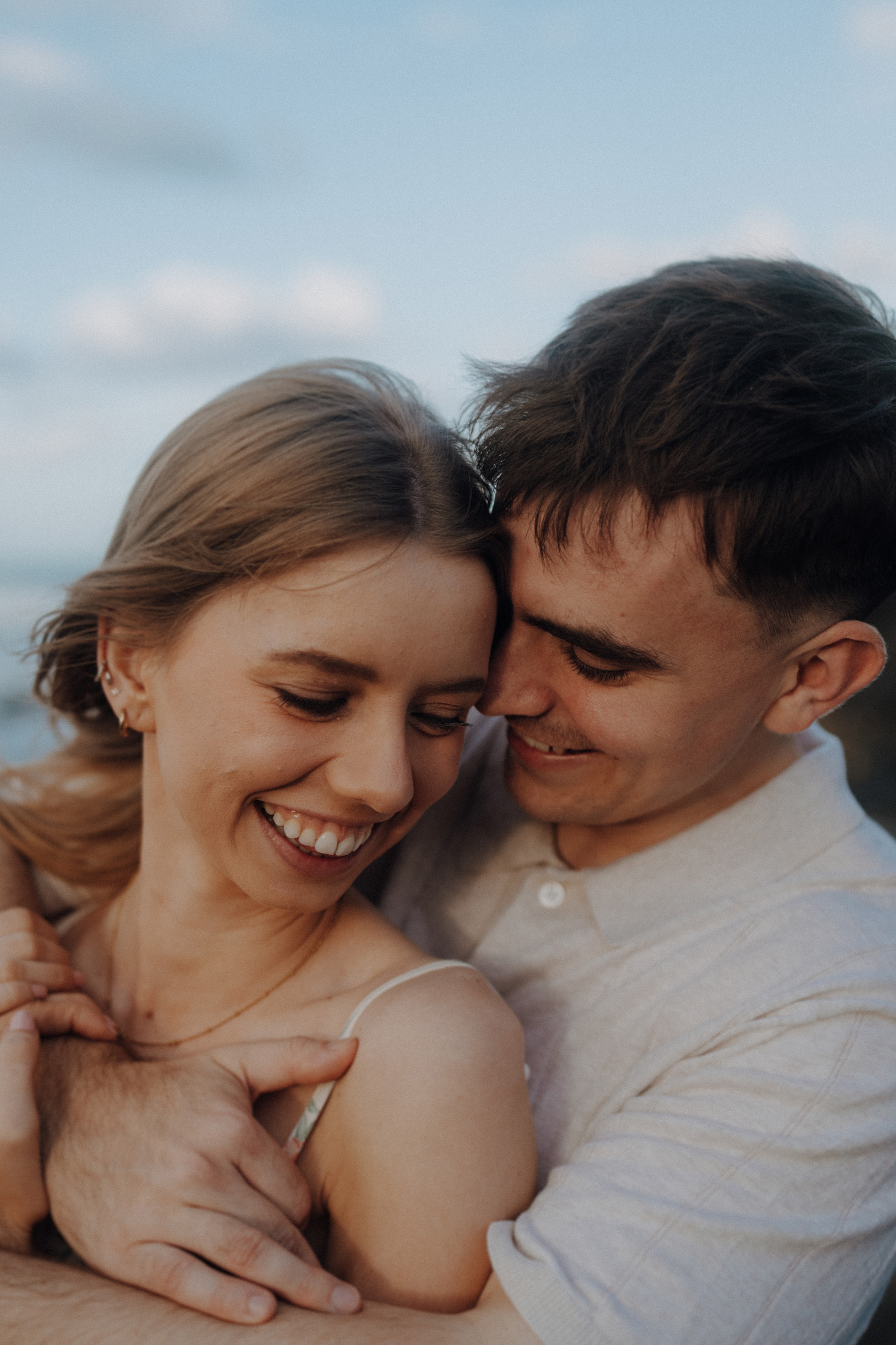 A couple embraces outdoors, both smiling. The woman is in front, while the man stands behind her, resting his head near hers. The sky is visible in the background.